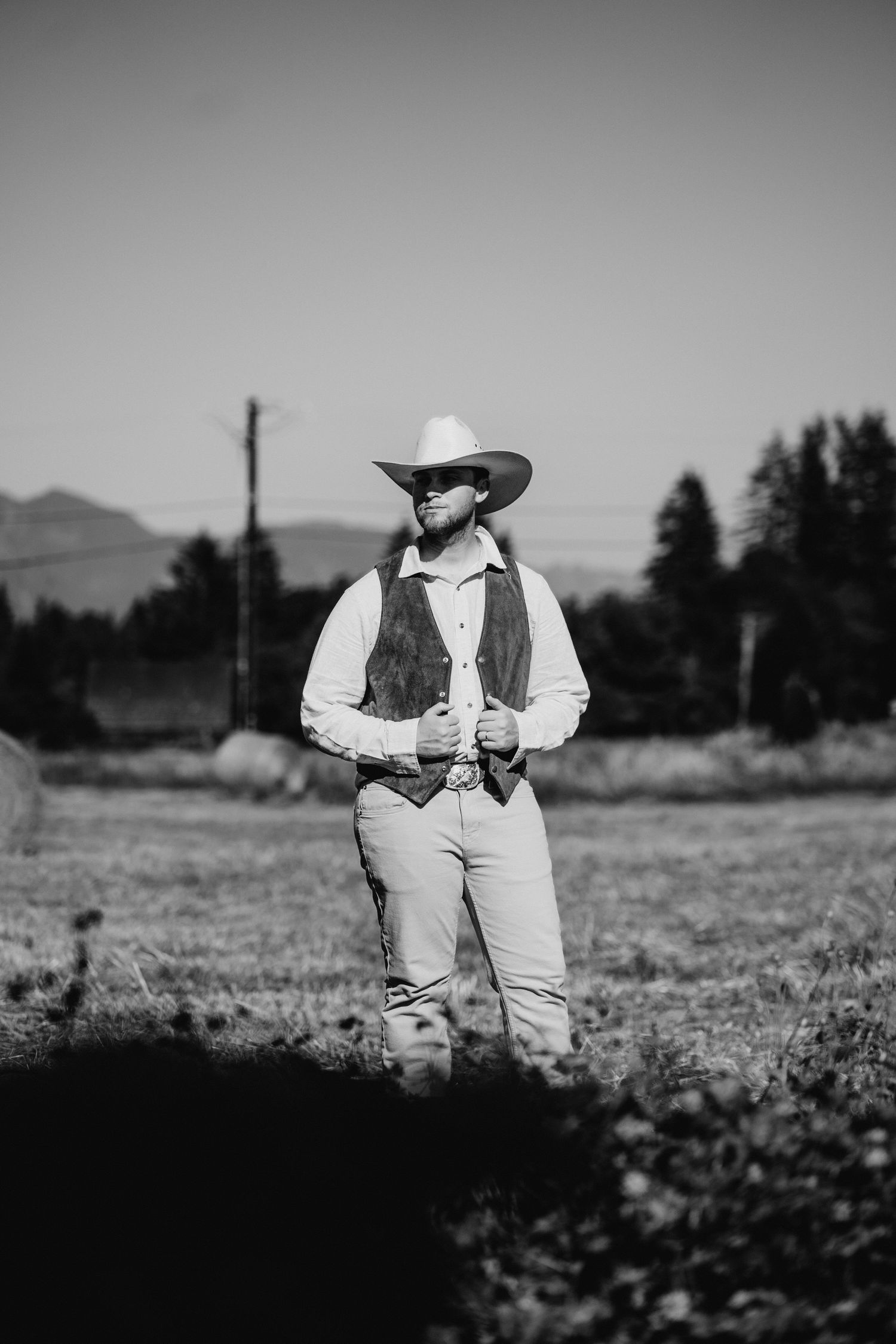 Black and white portrait of groom in cowboy hat and suede vest in farm field.