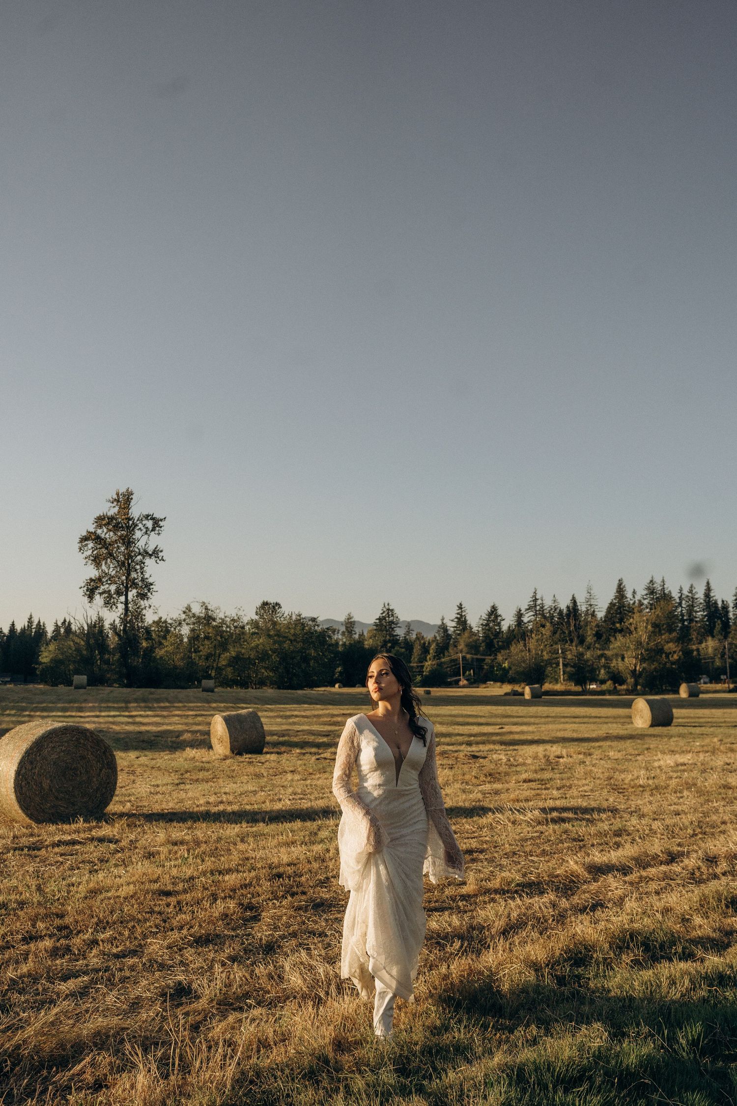 Bride in white lace gown walks through a golden field with hay bales at sunset.