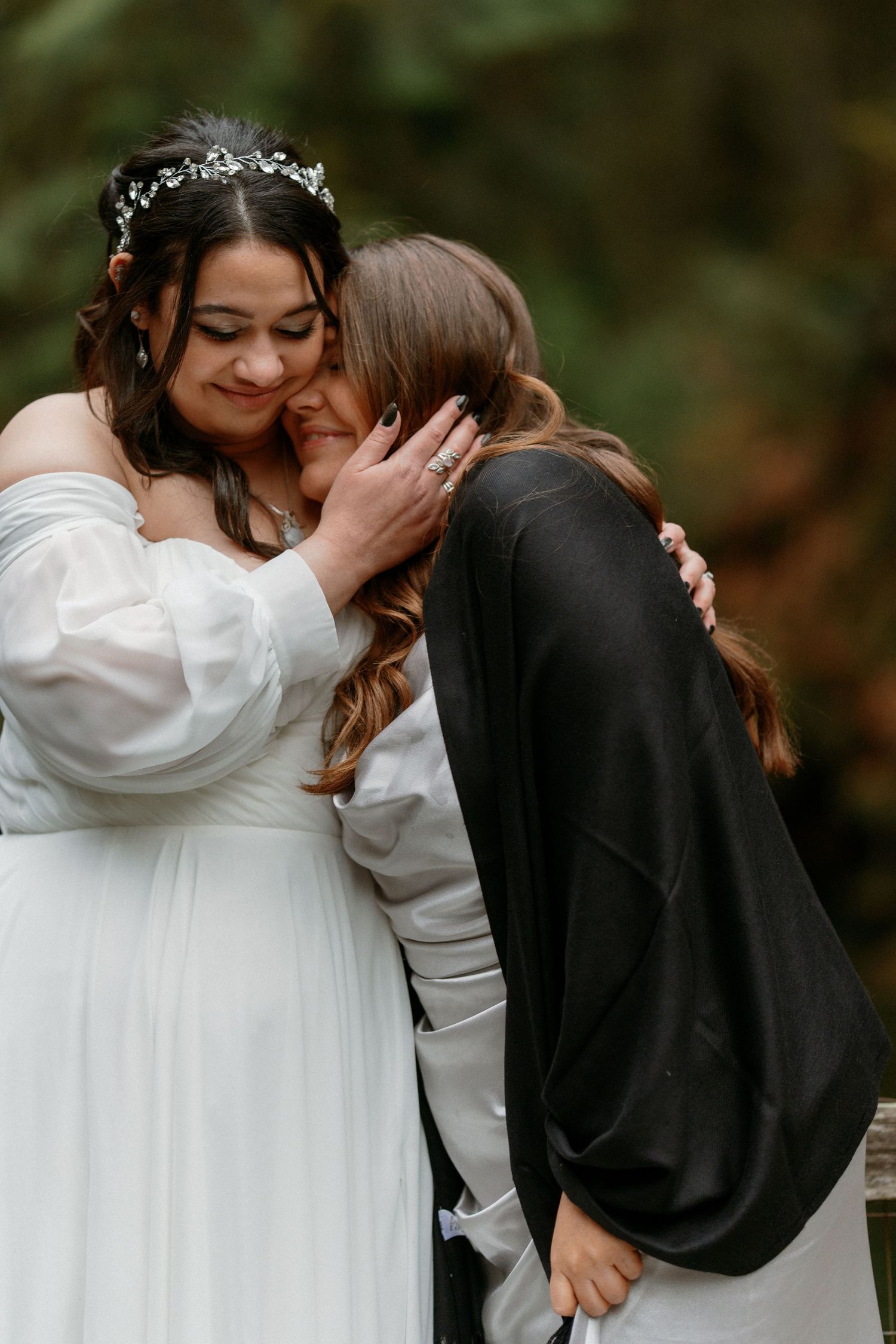 Two women embracing bride in white wedding gown and floral crown outdoors.