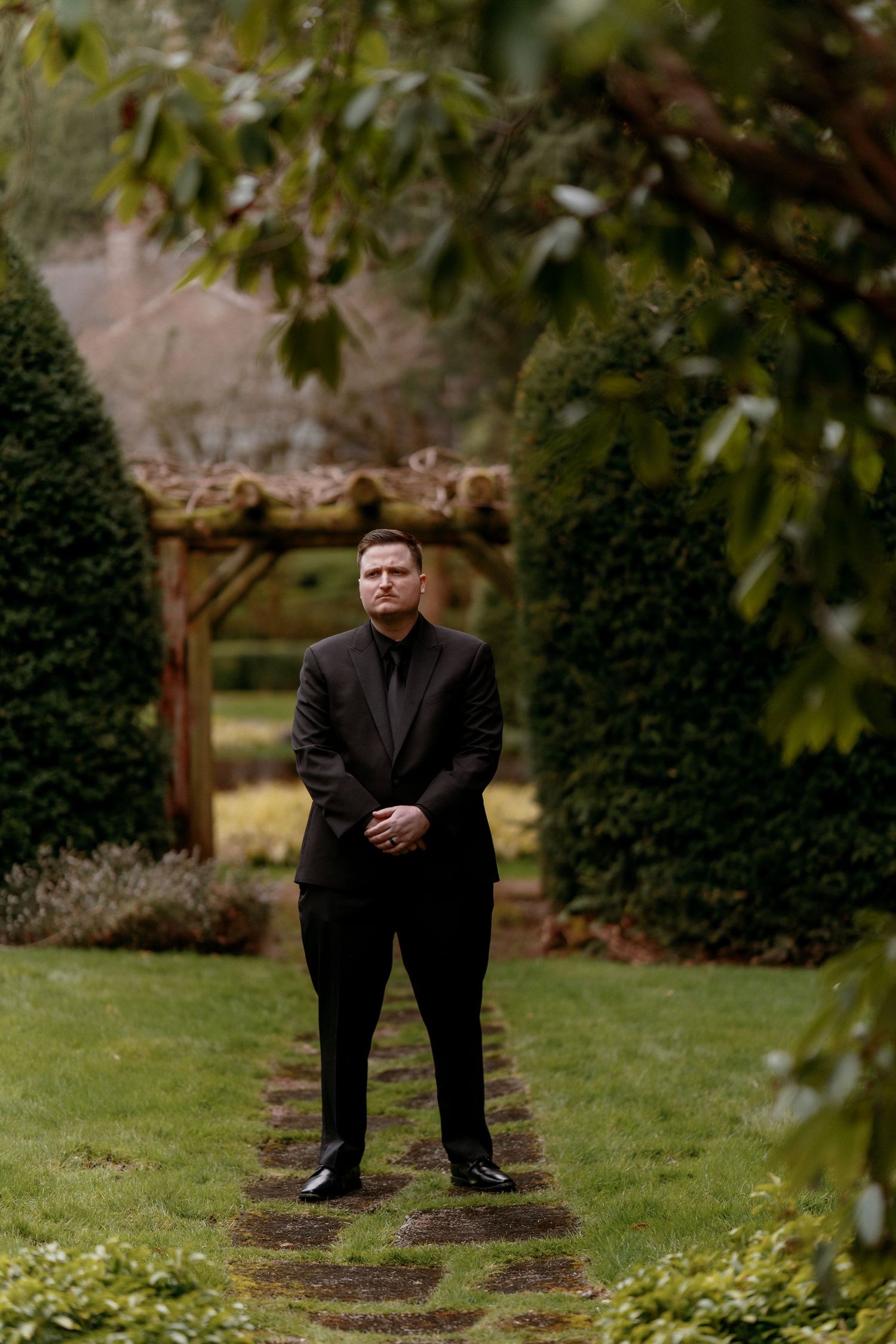 Groom in all-black suit standing on garden path framed by lush hedges and a wooden arbor.