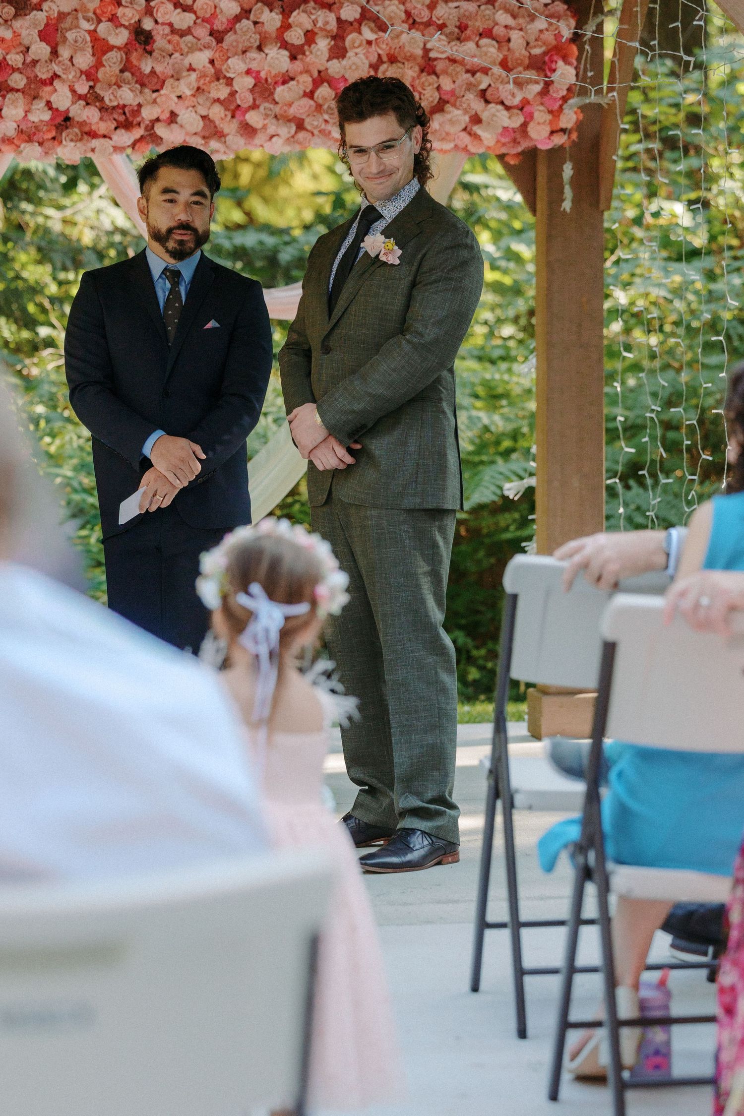 Groom in gray suit watches flower girl walk down aisle at outdoor woodland wedding ceremony.