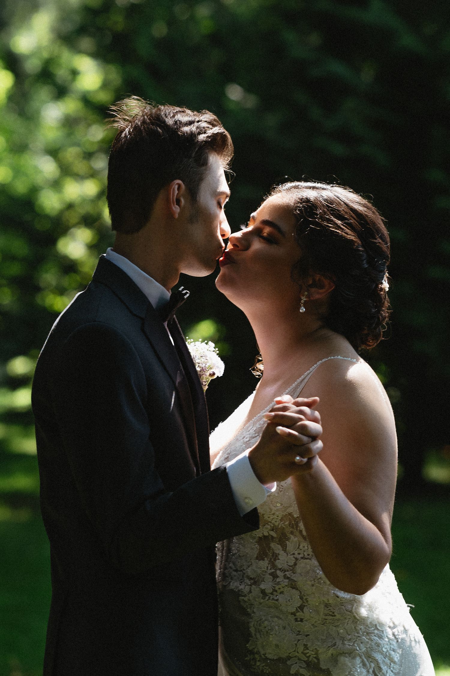 Bride and groom share a romantic kiss outdoors with lush green trees in the background.