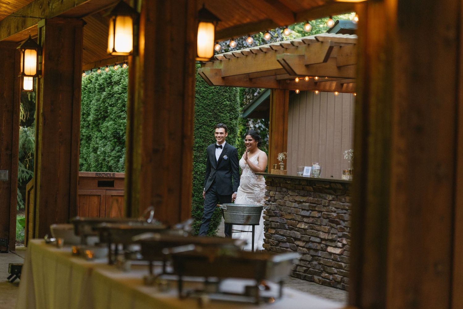 Bride and groom stand together at rustic outdoor wedding venue with string lights and buffet.