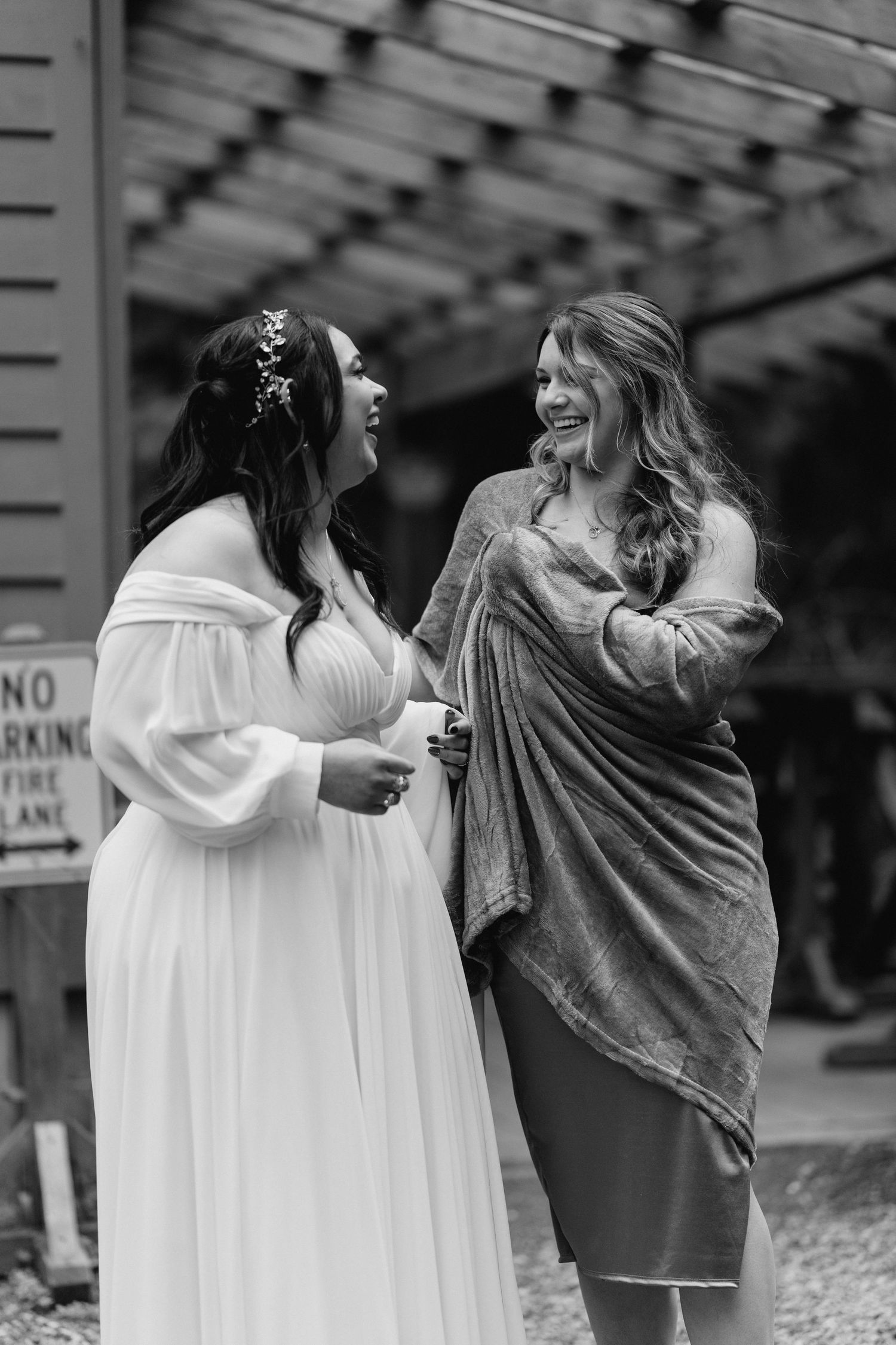 Bride and bridesmaid laugh together joyfully outside rustic wedding venue in candid photo.