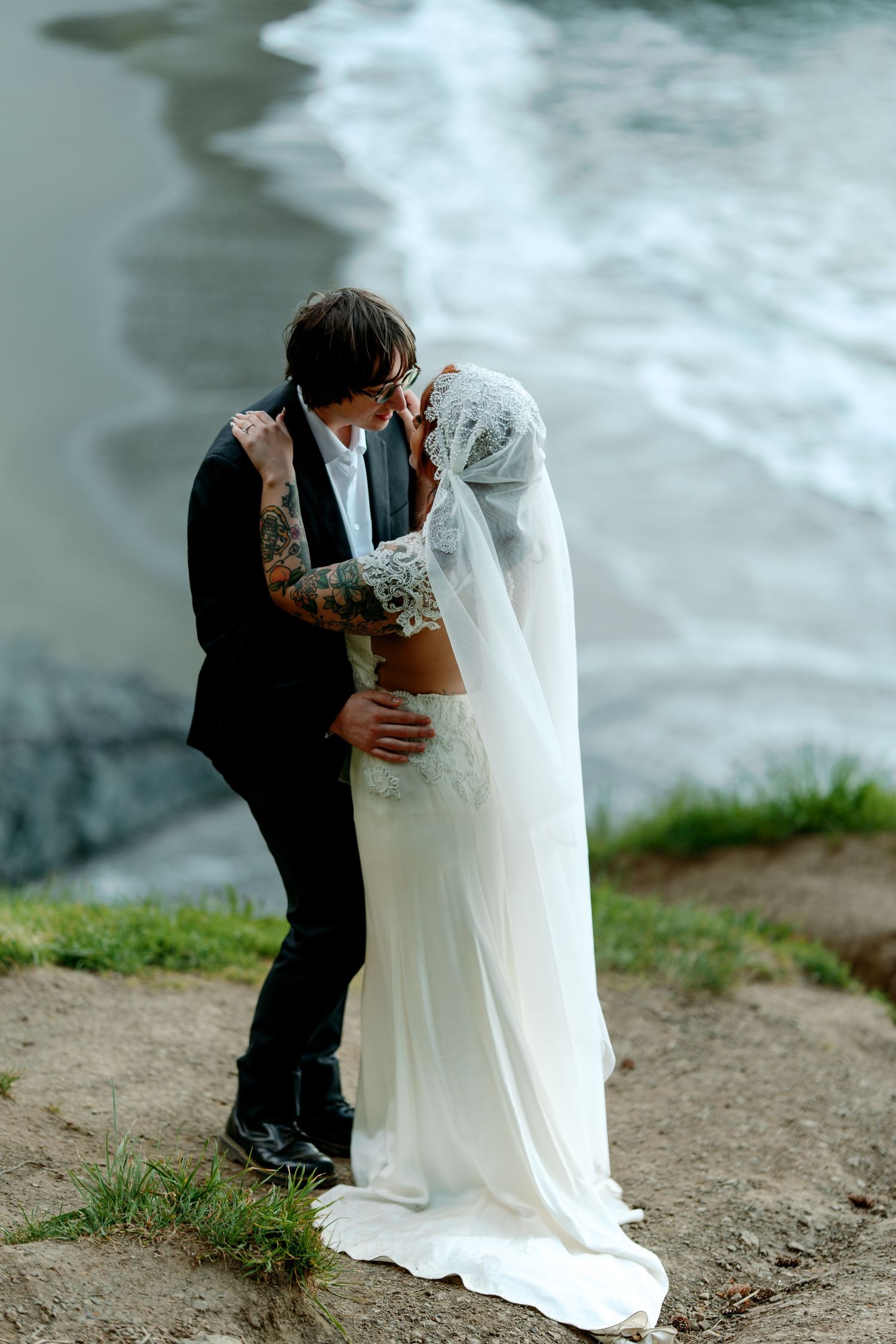 Tattooed bride and groom share a romantic kiss on a coastal cliff with ocean waves in the background.