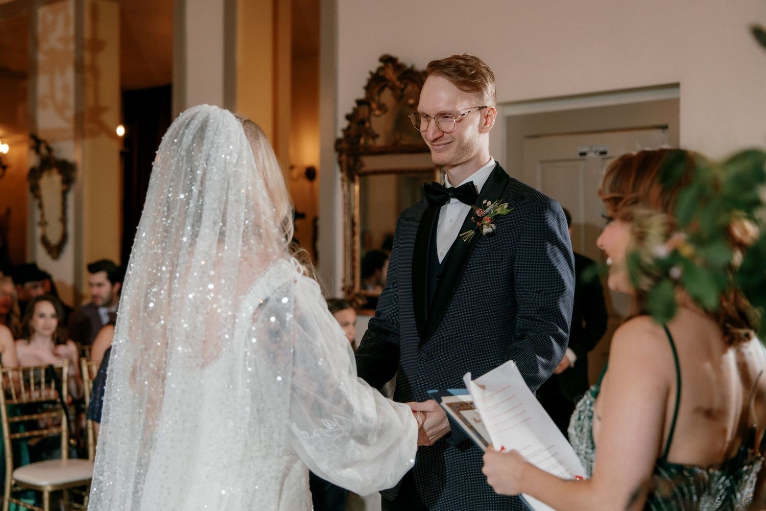 Bride and groom exchange vows indoors, smiling at each other while holding hands during their wedding ceremony.