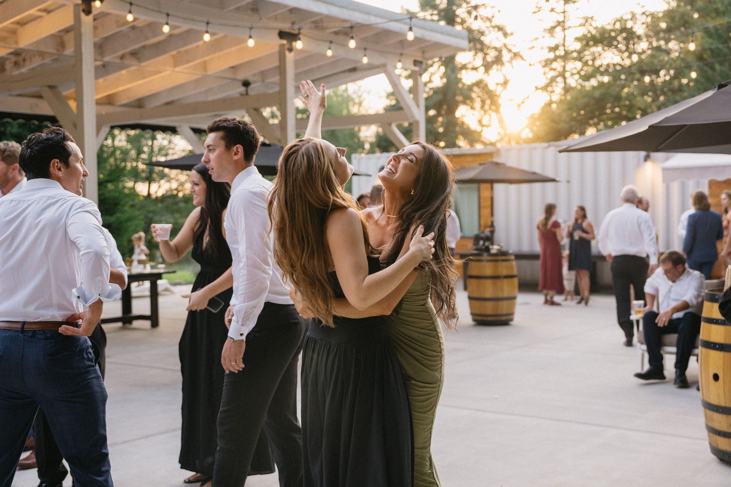 Wedding guests embracing and dancing at golden hour outdoor reception with string lights.