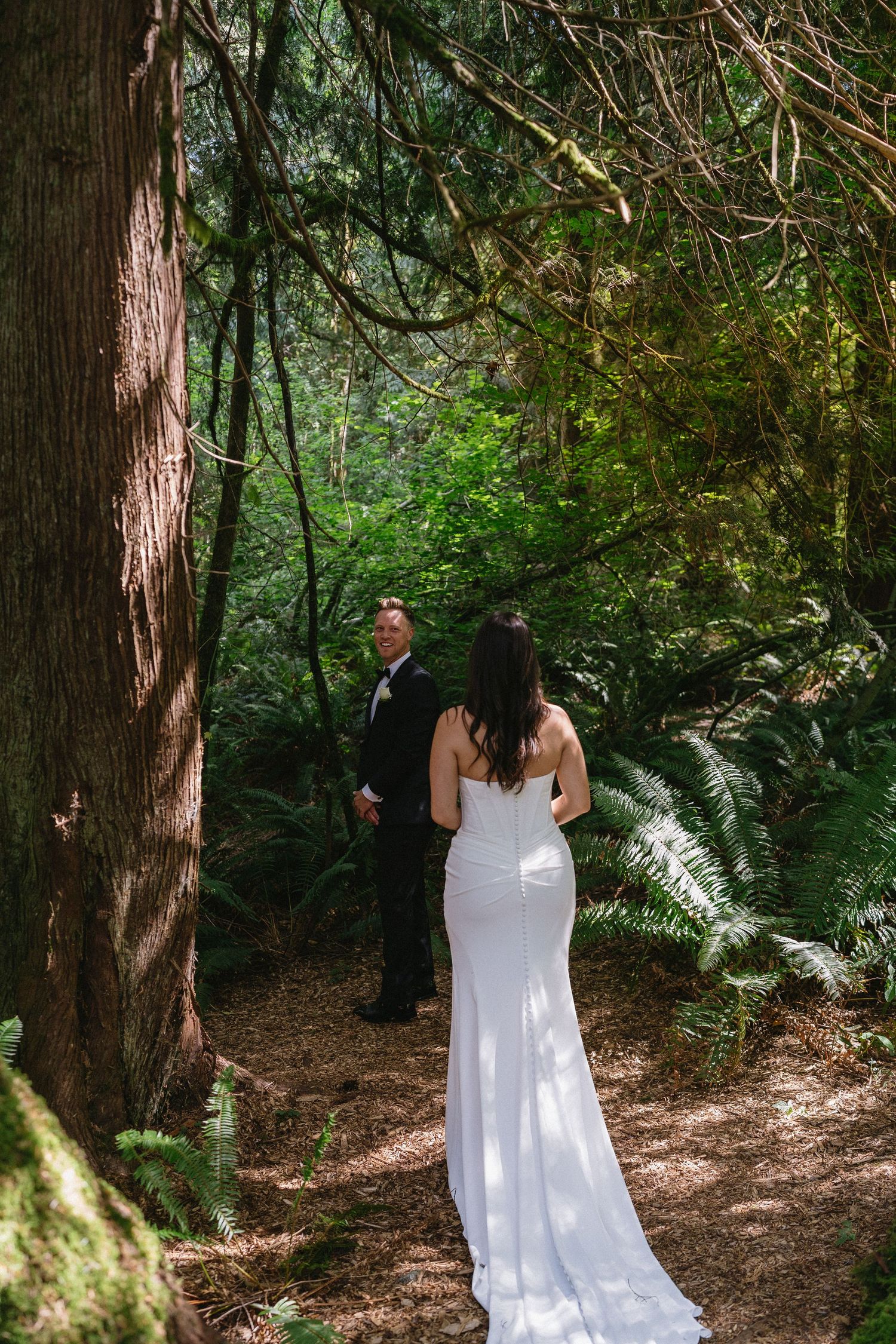Bride walks toward groom in black tuxedo on sunlit forest trail.