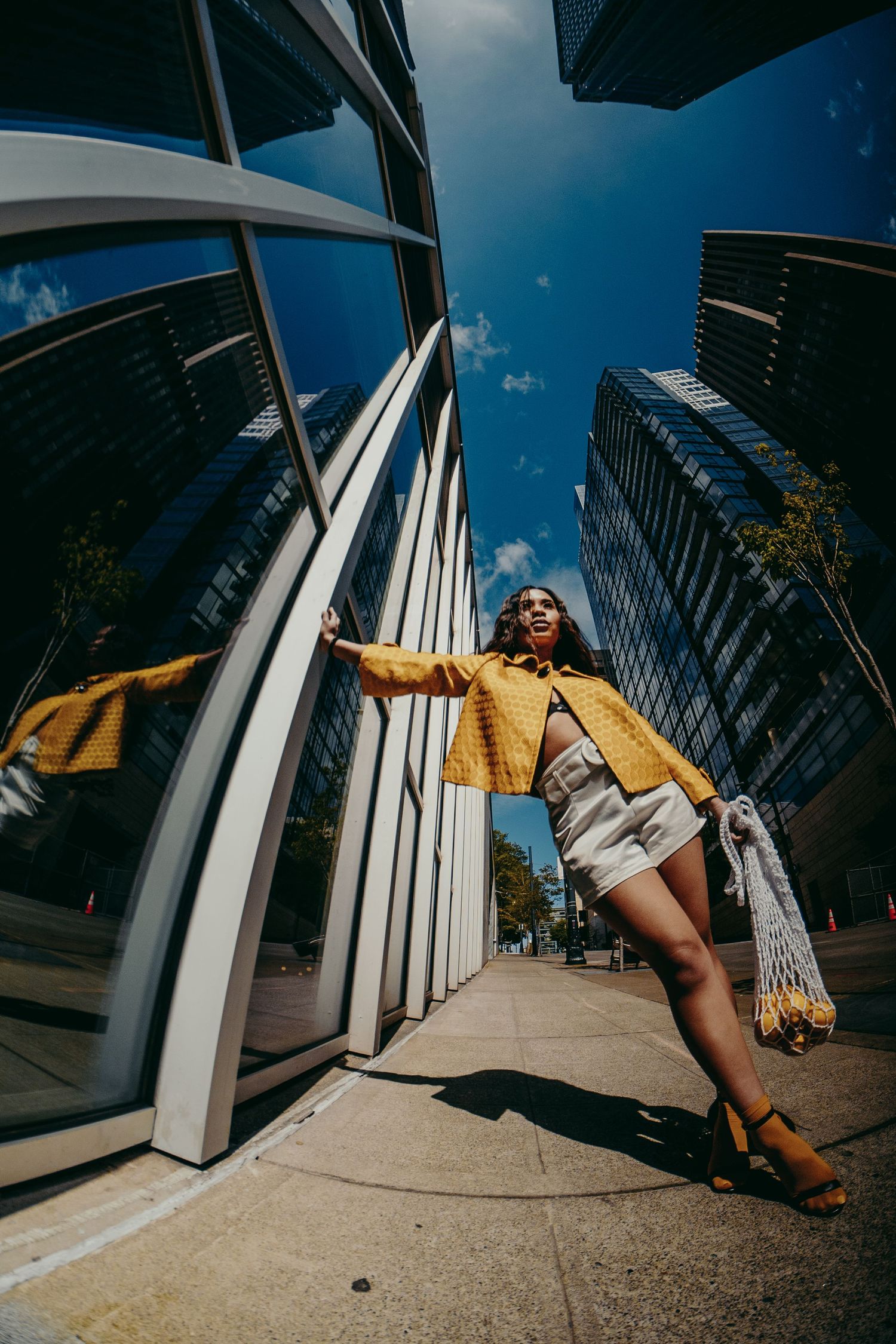 Woman in yellow jacket and white shorts poses boldly between curved glass skyscraper walls using fisheye lens.