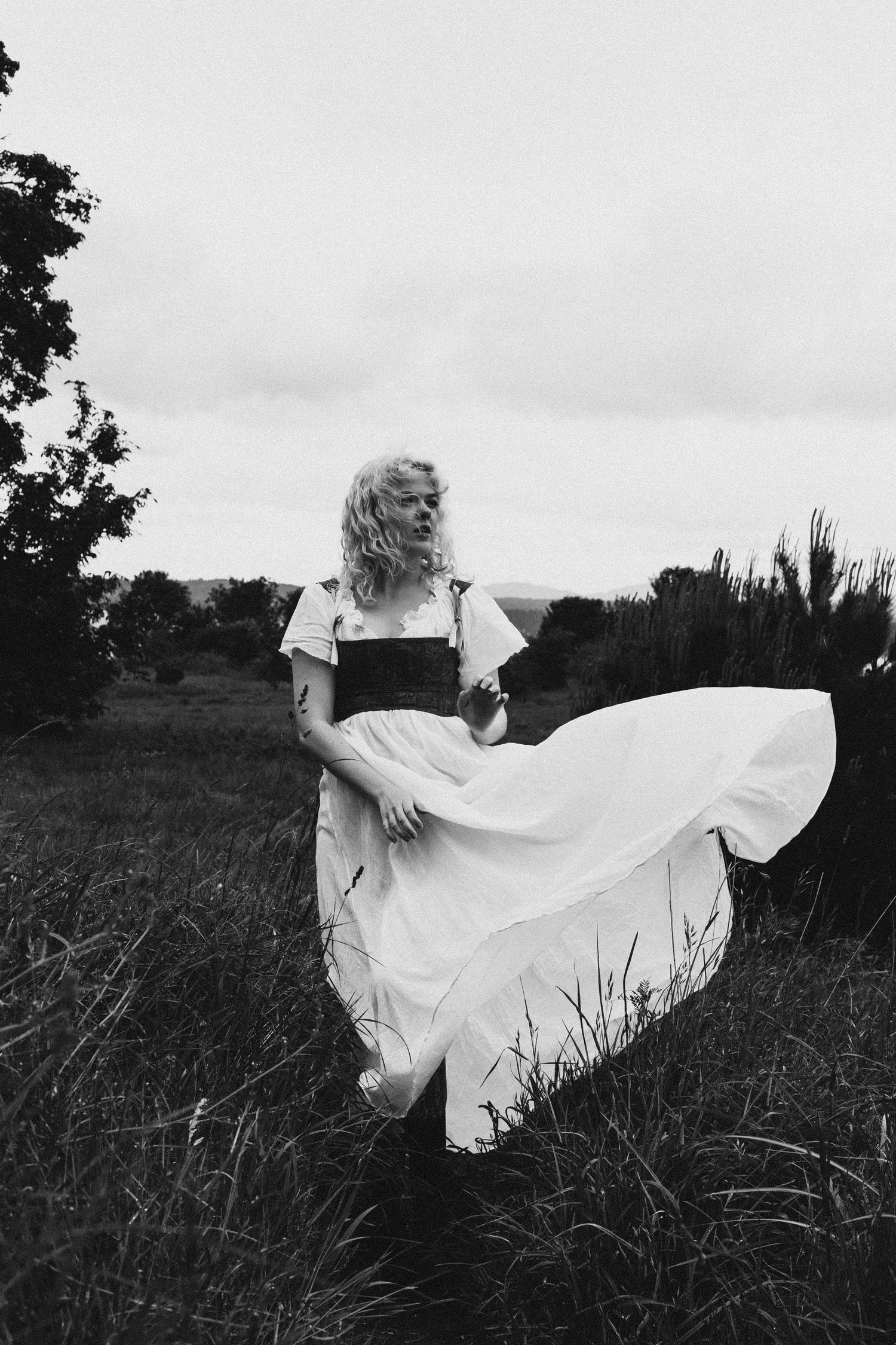 A blonde woman in a flowing white dress and black corset stands in a grassy field in a moody black and white photo.