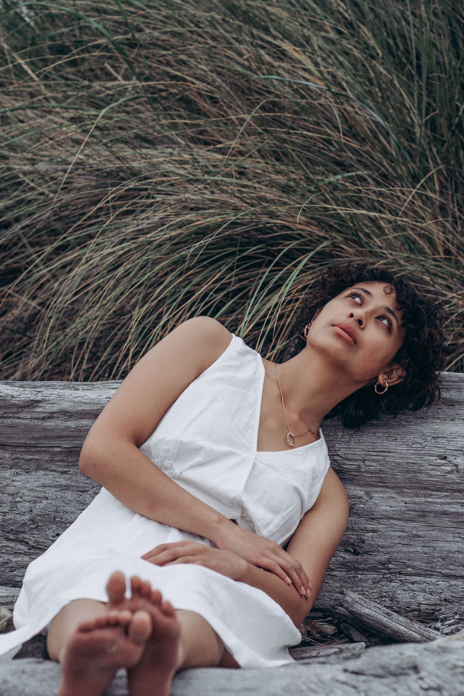 Barefoot woman in white dress sits against driftwood gazing up surrounded by tall grass.