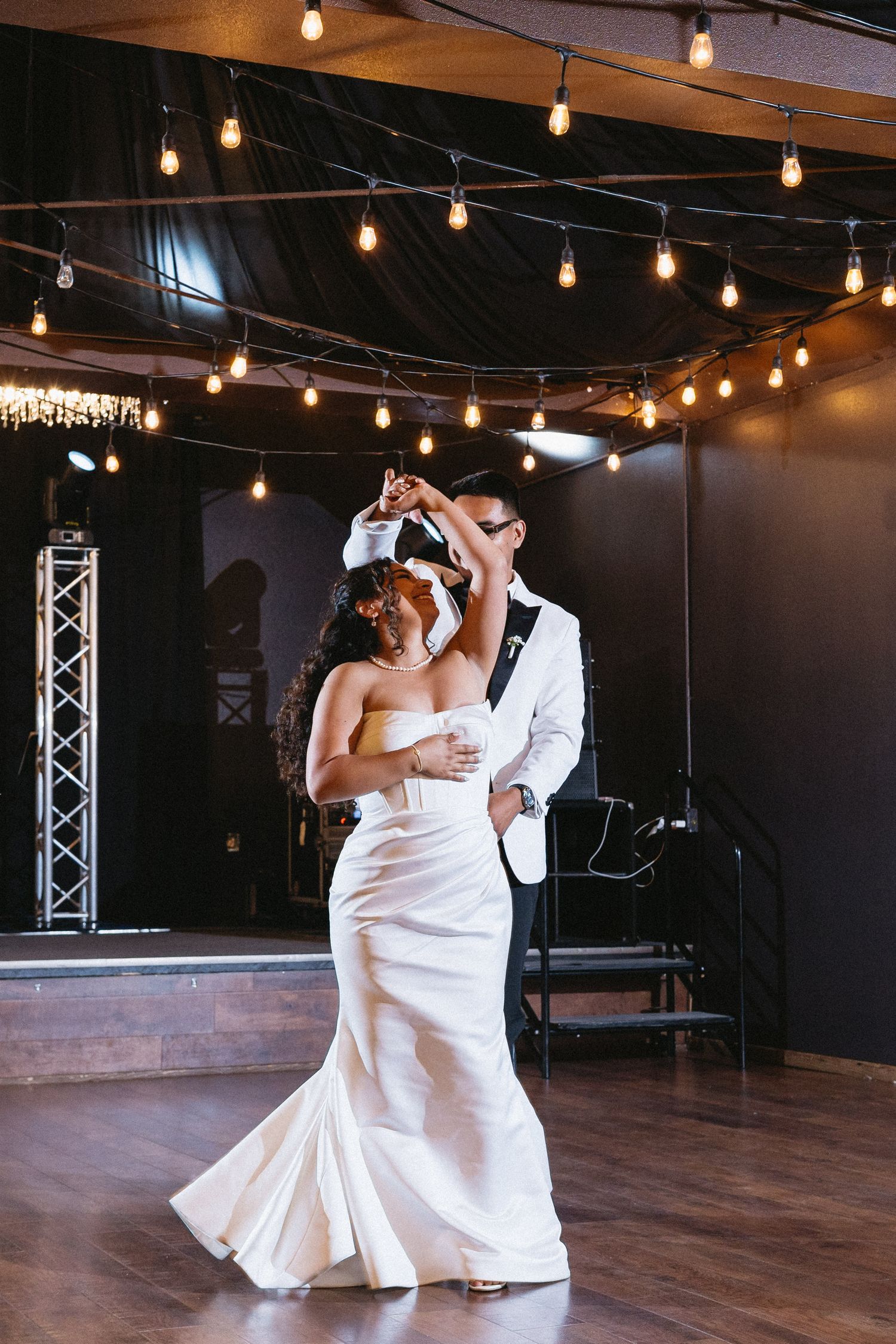 Groom twirls bride in elegant white gown during first dance under glowing Edison bulb string lights.