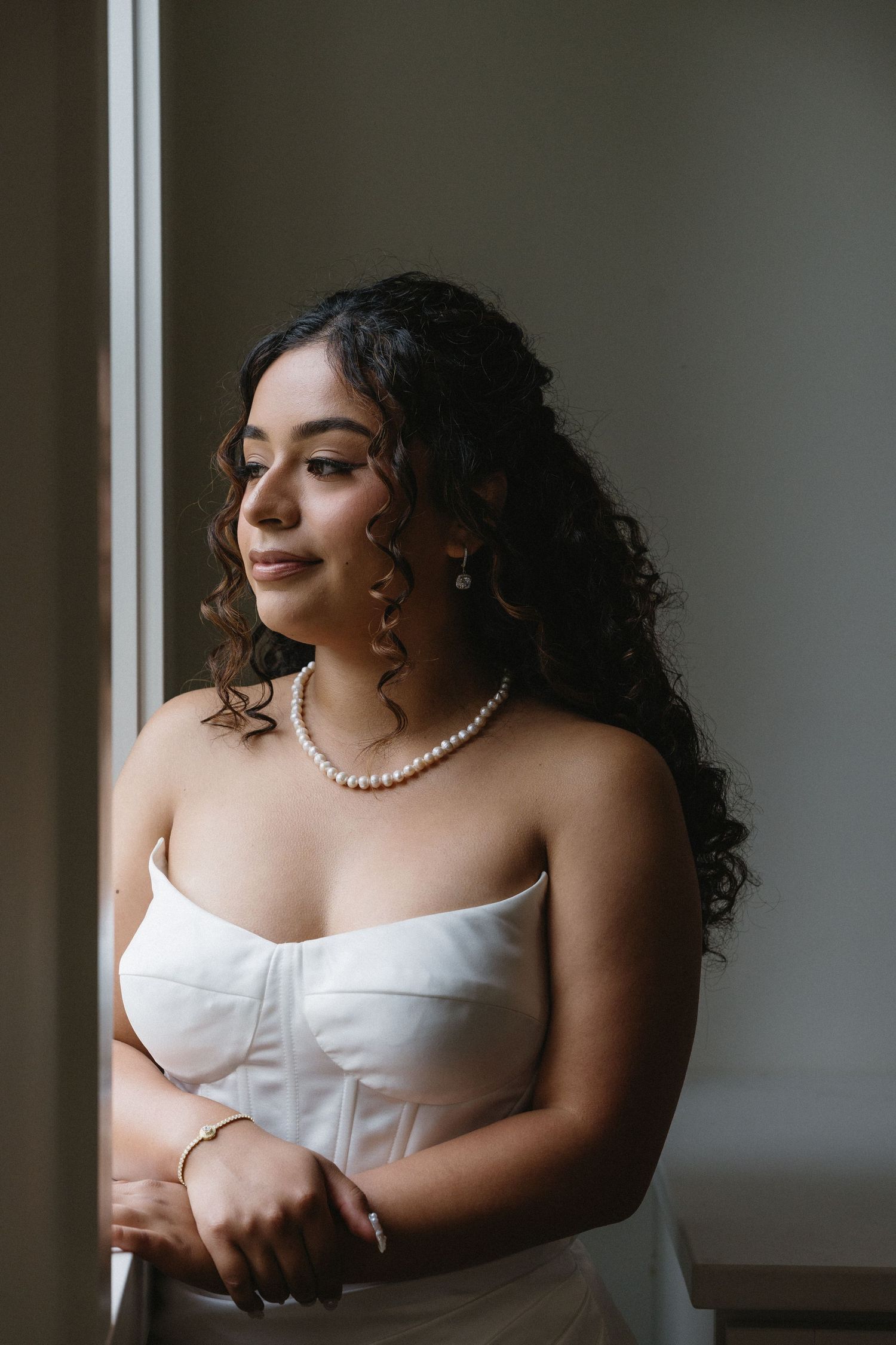 Elegant bride in strapless white gown wearing pearl necklace and earrings gazing out window.