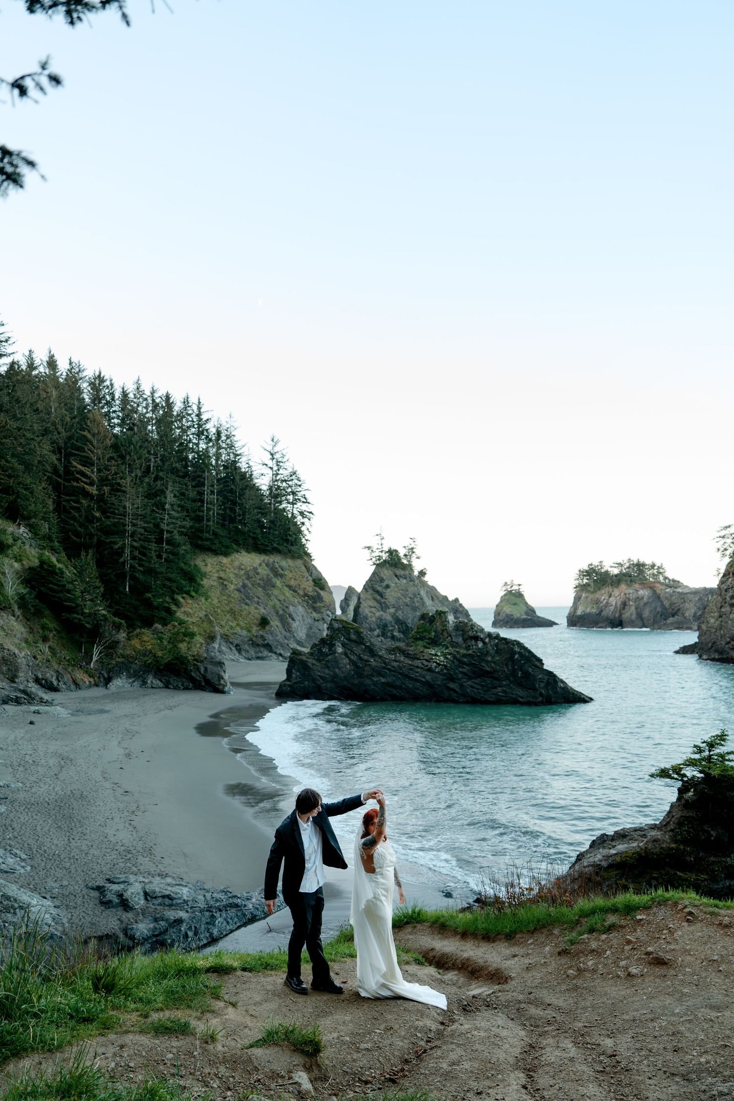 Bride and groom stand on coastal cliffside overlooking a dramatic rocky beach during elopement ceremony.
