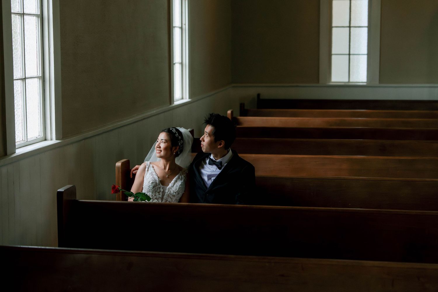 Bride and groom sitting together in wooden church pews near sunlit windows.