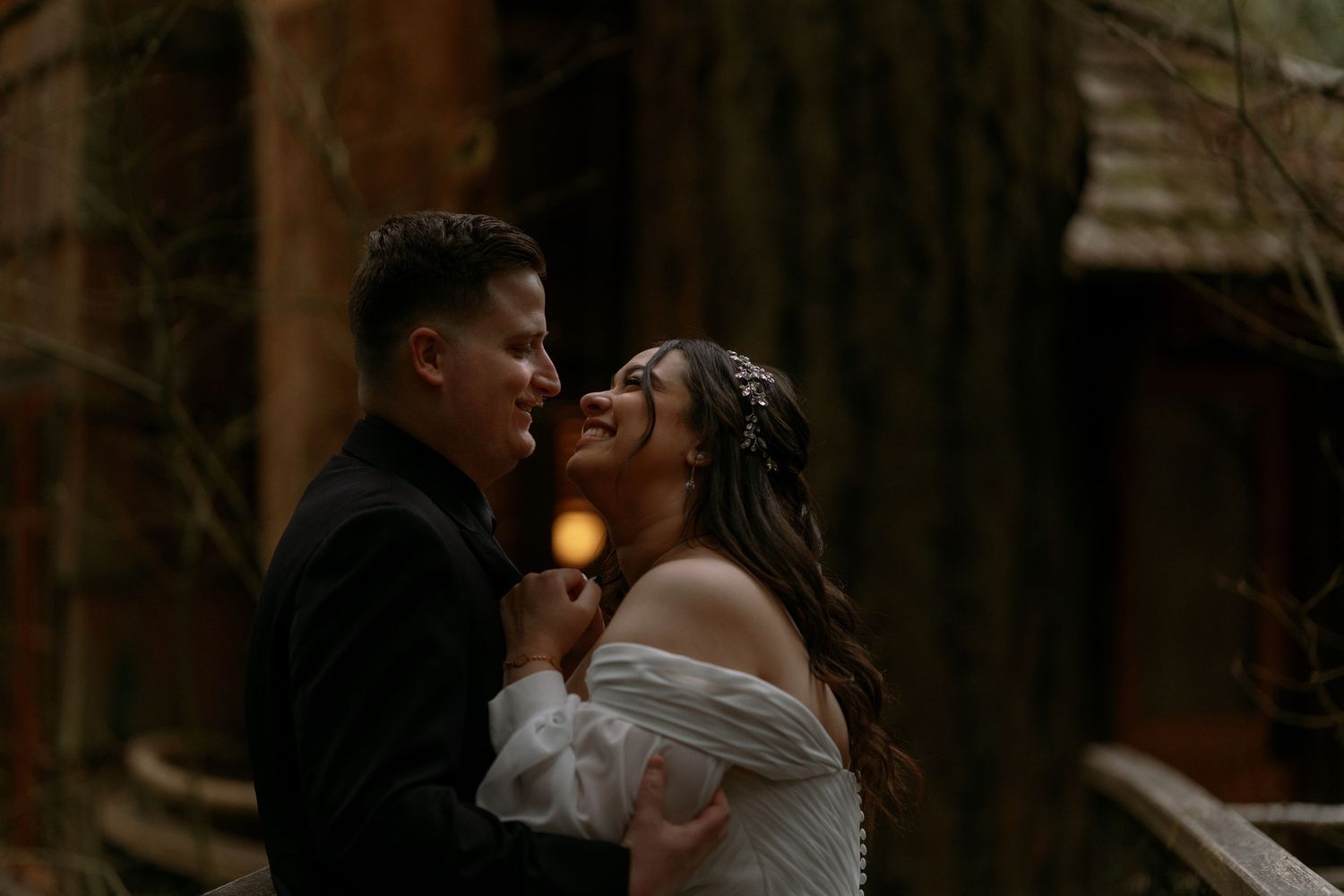 Bride laughs joyfully as groom gazes at her lovingly during romantic twilight wedding portraits in woods.