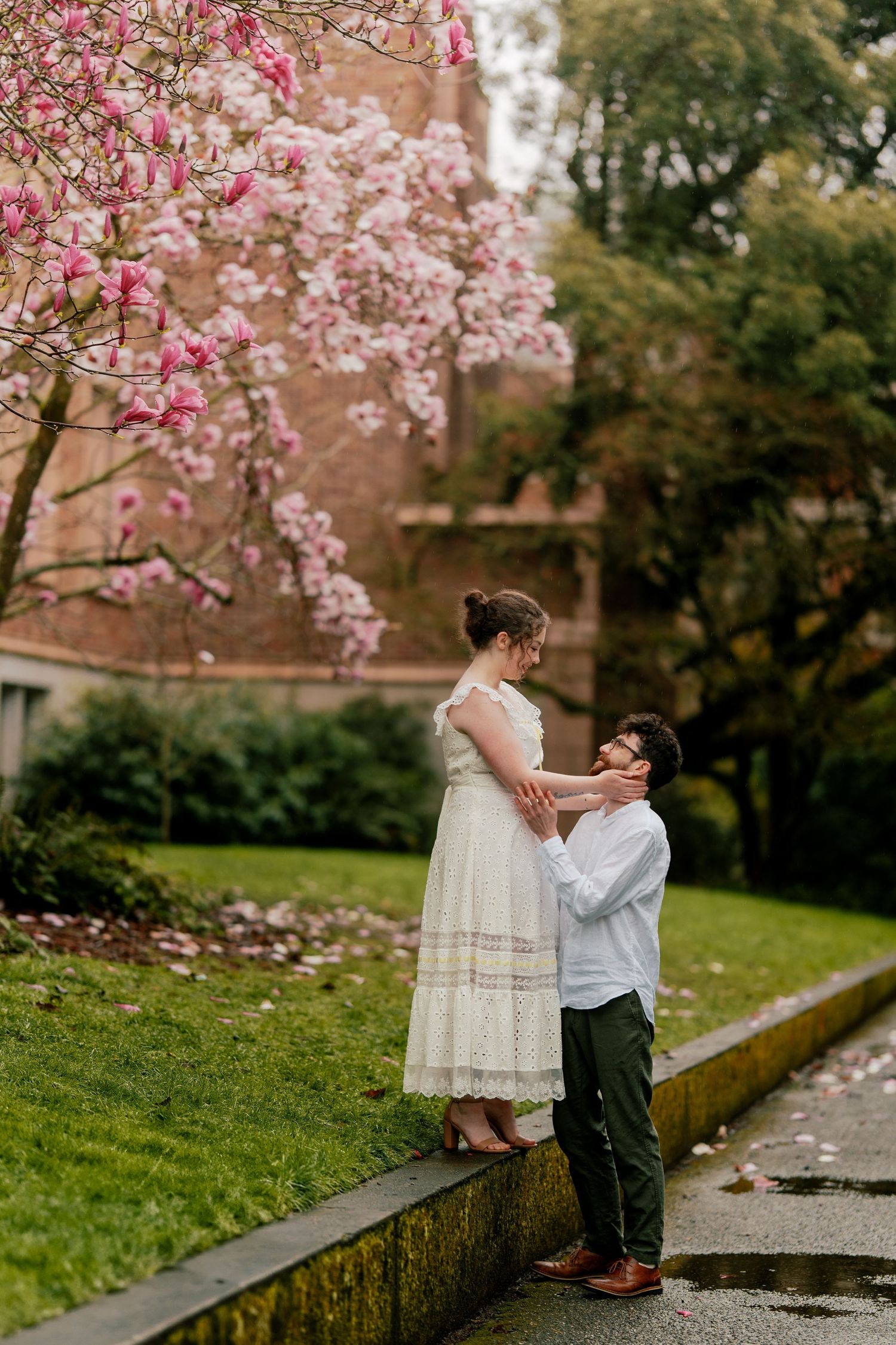 Couple standing together under pink magnolia blossoms near a brick building in a rainy garden.