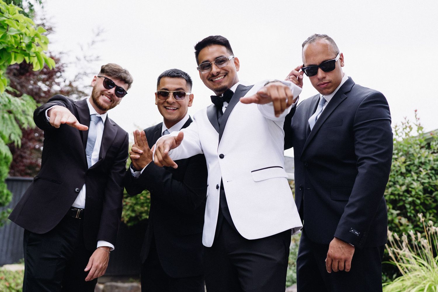 Four groomsmen in black suits with sunglasses posing confidently outdoors before wedding ceremony.