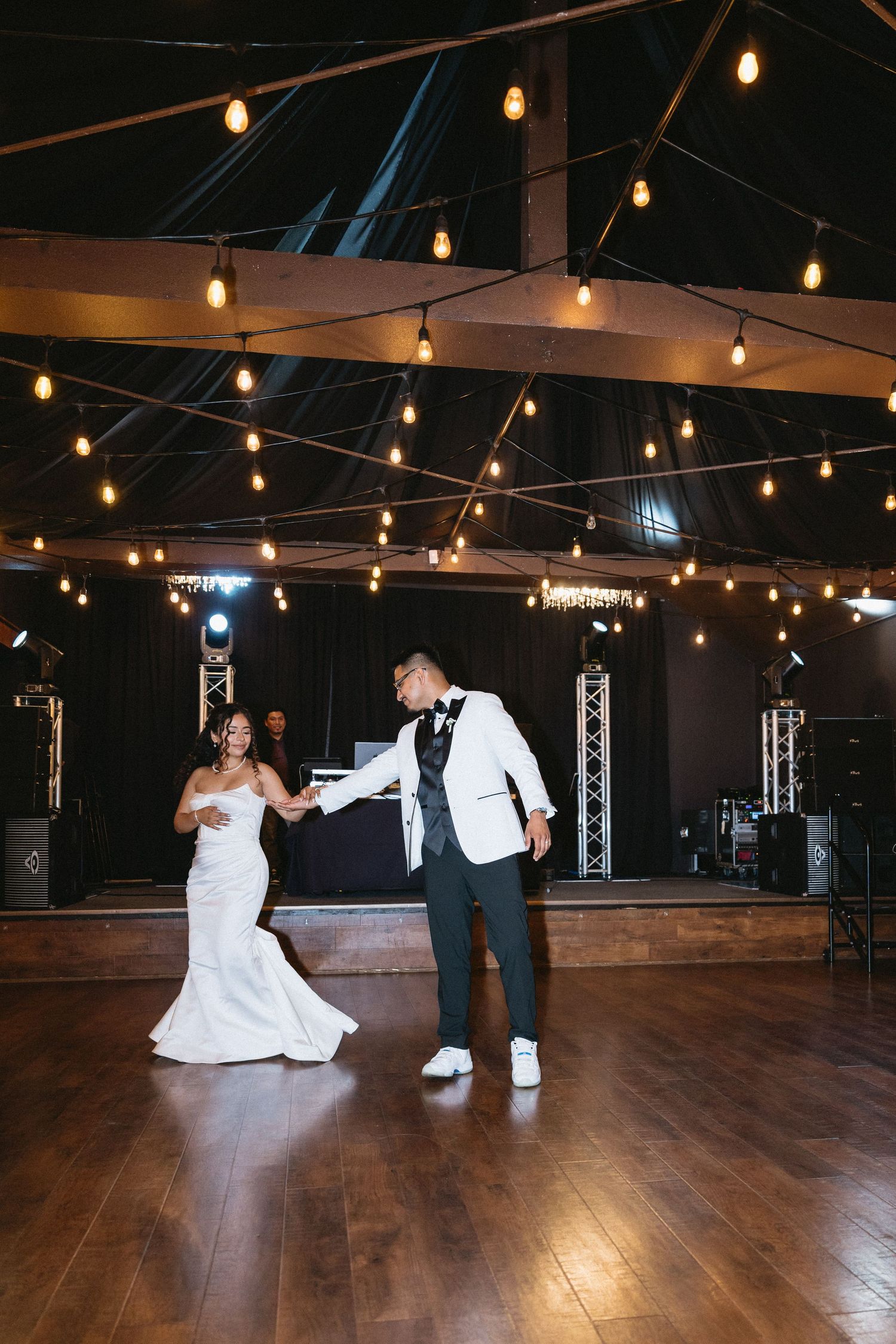 Bride and groom share their first dance on a wooden floor under warm string lights at a wedding reception.