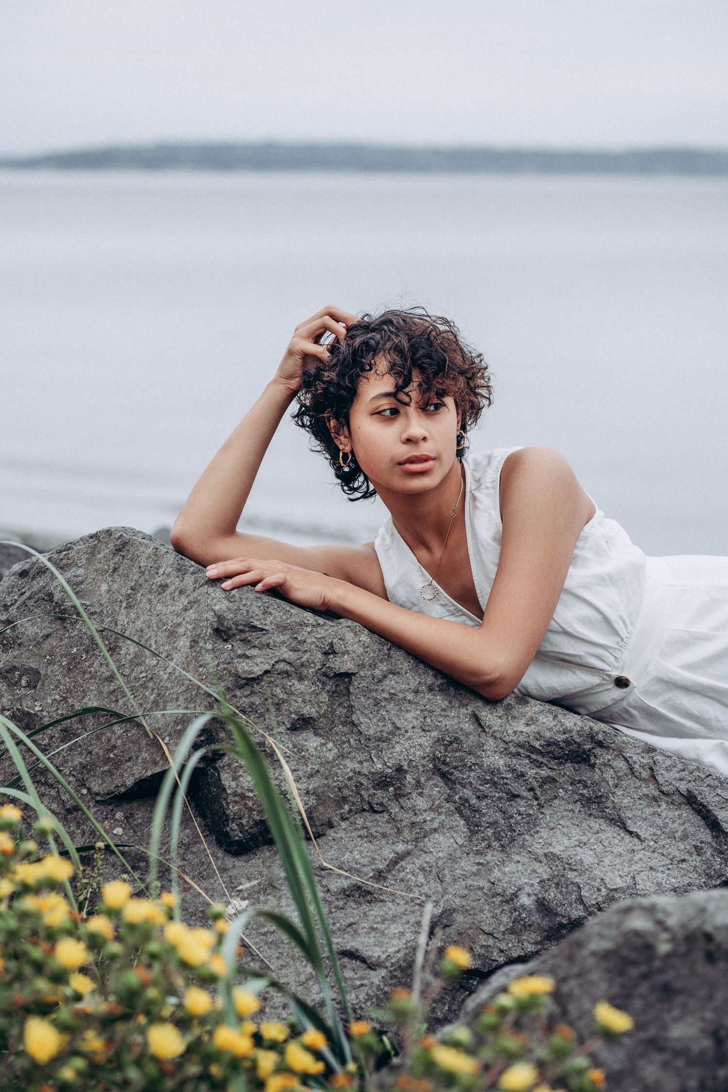 Woman in white dress leans on rock by water with yellow wildflowers in foreground.