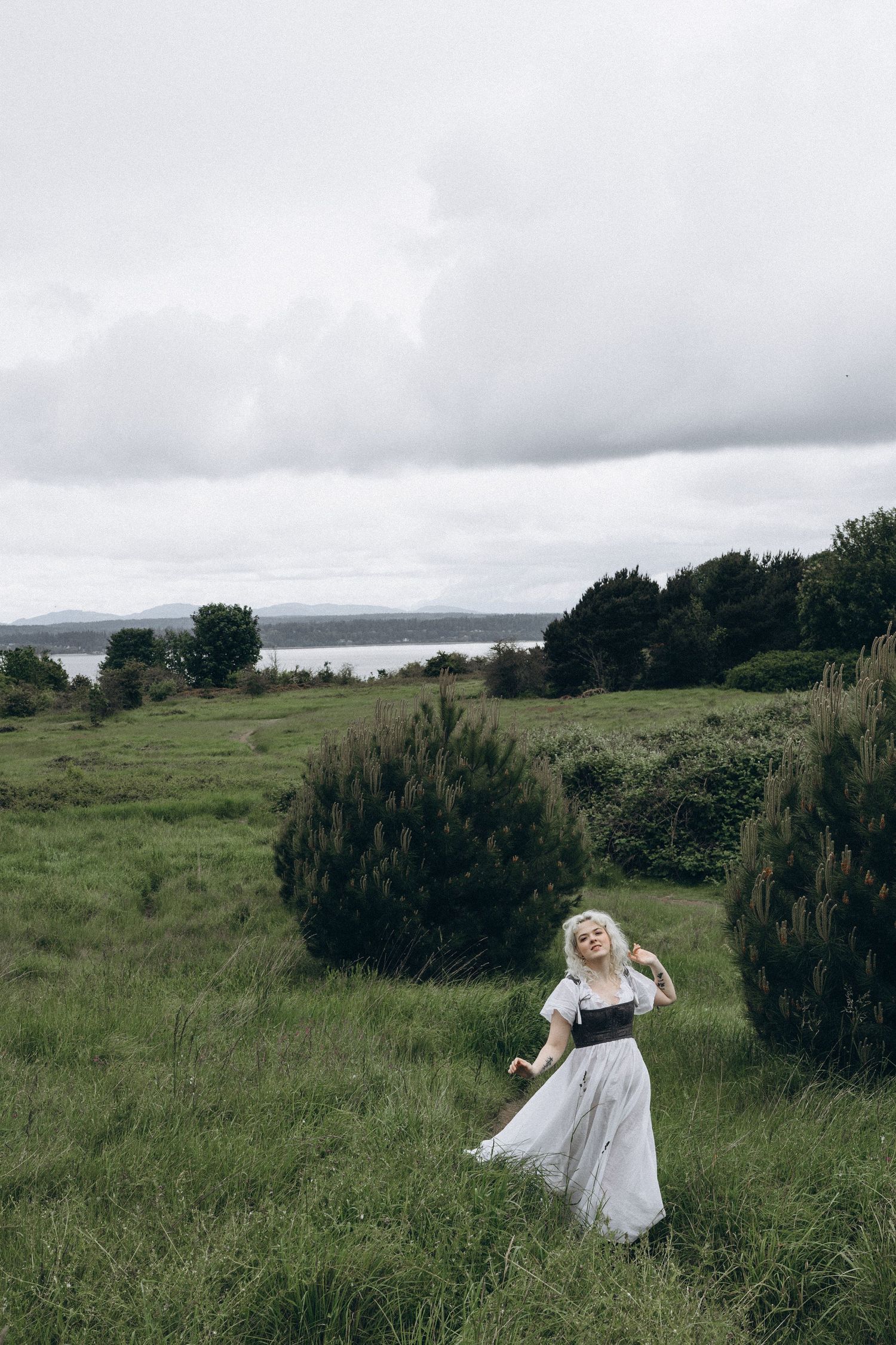 A blonde woman in a white dress and black corset walks through a lush green field with a lake in the background.
