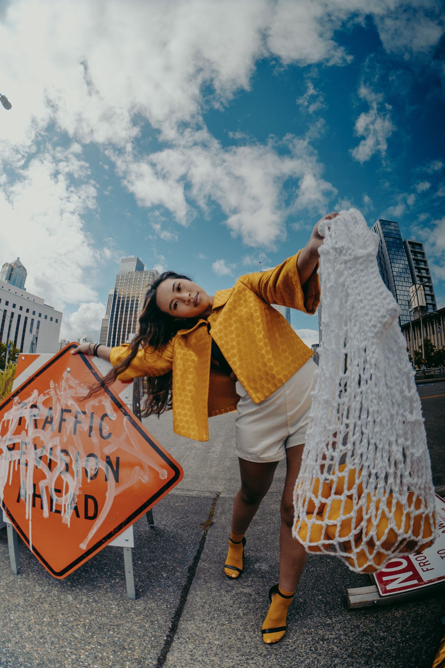 Woman in yellow jacket holds a mesh bag near a traffic sign with city skyscrapers in the background.