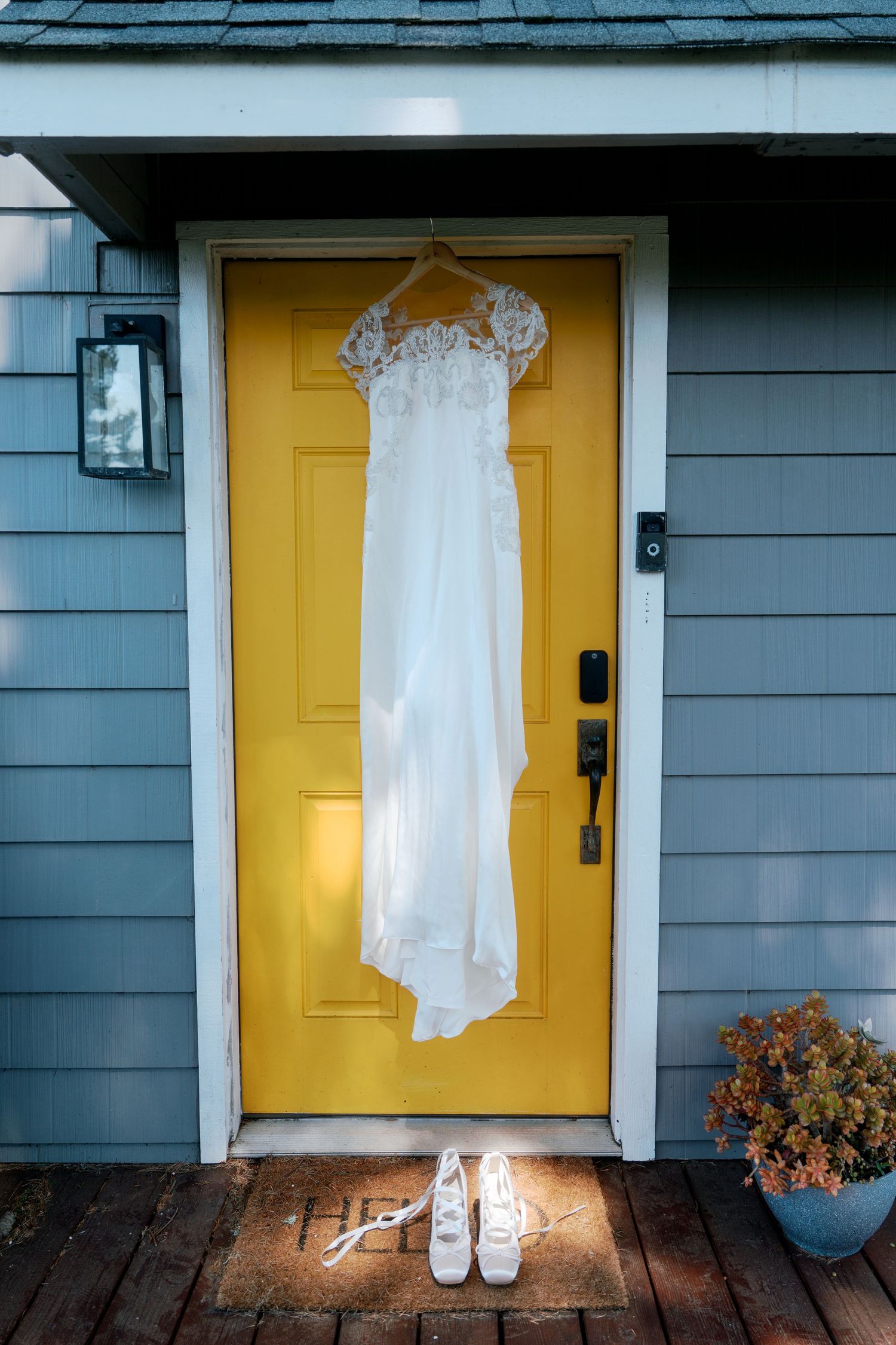 Elegant lace wedding dress hangs on bright yellow front door with white bridal sneakers placed below on porch.