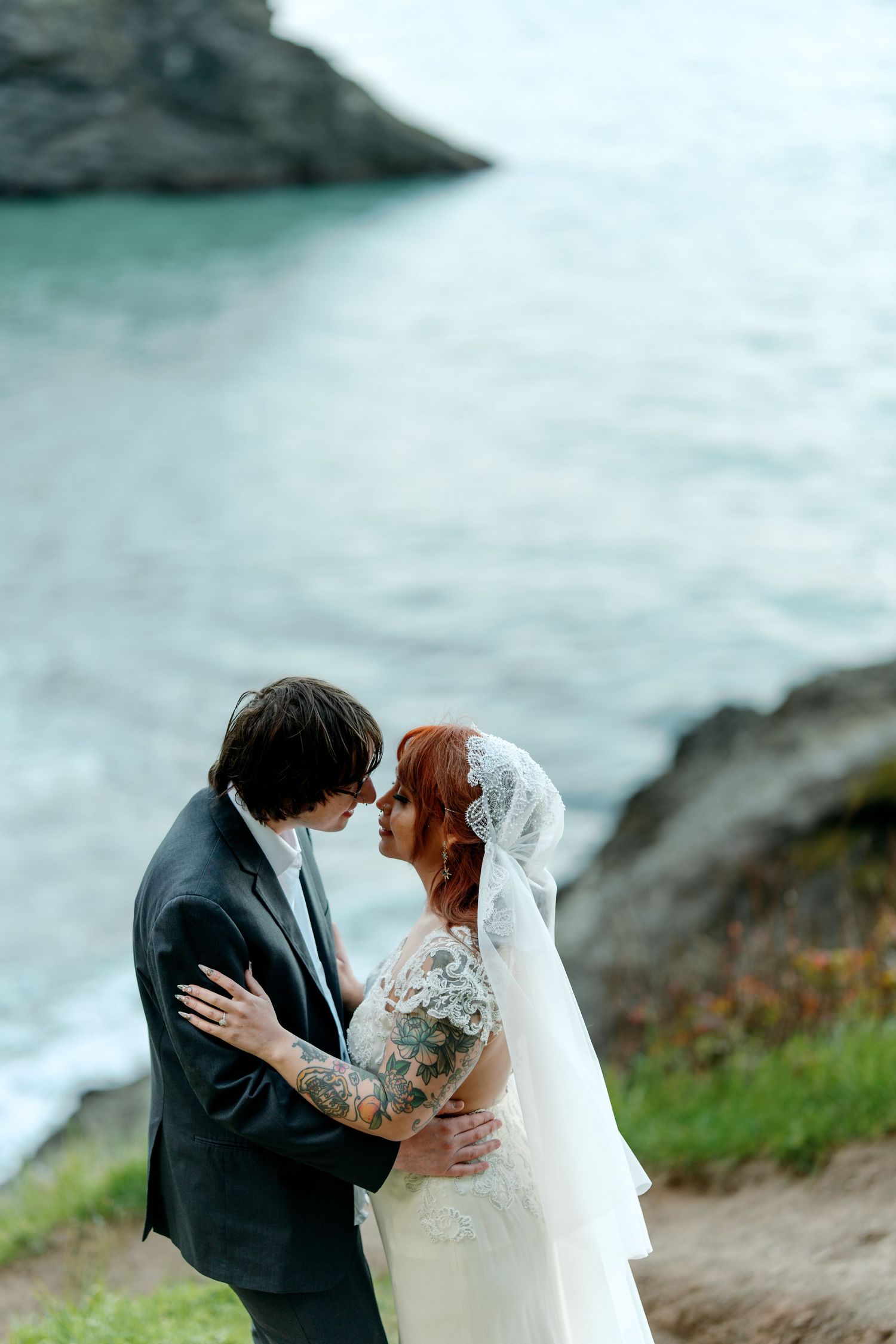 Bride with red hair and tattoos and groom embrace intimately on a rocky ocean clifftop.