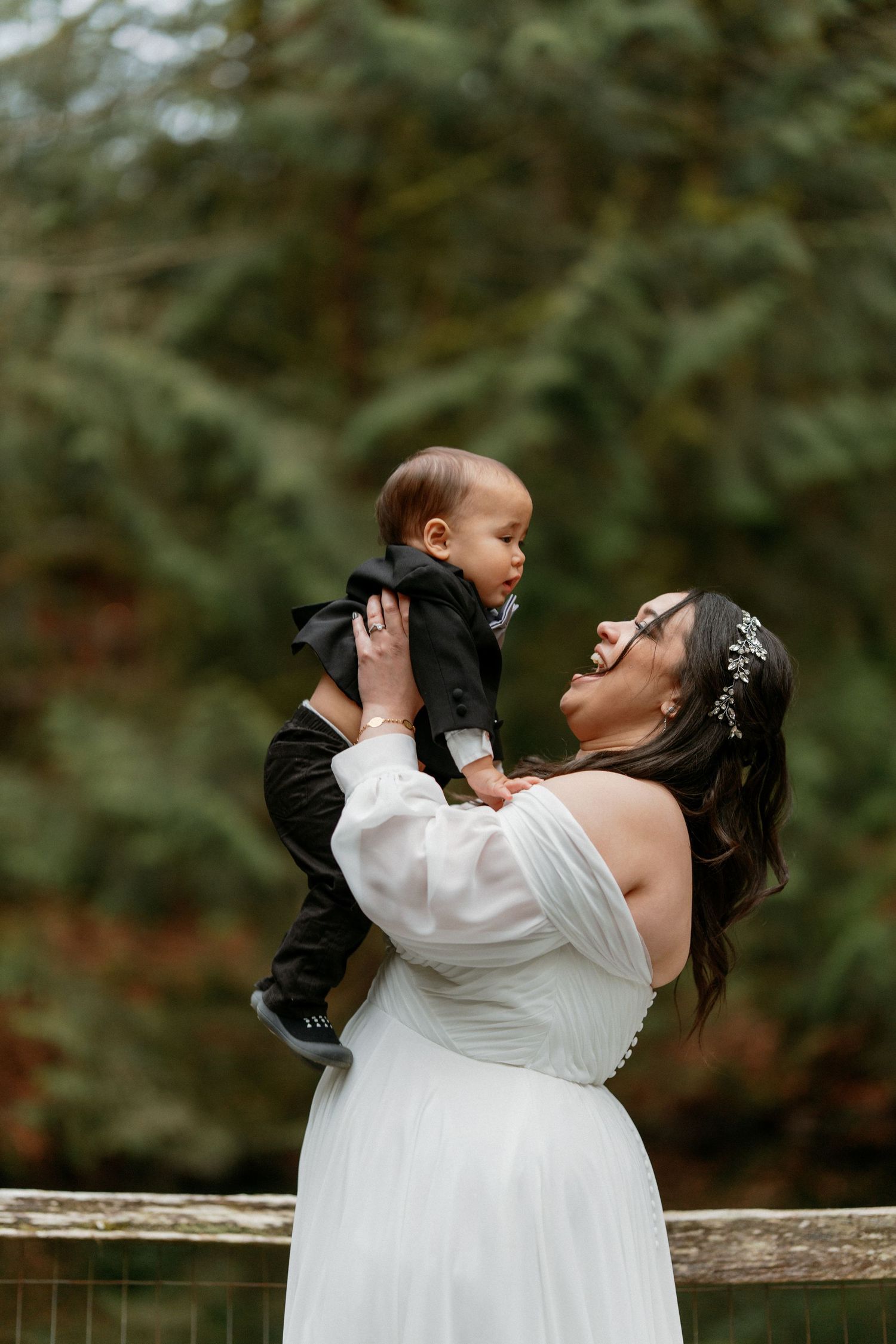 Bride in white gown holding smiling baby in black tuxedo outdoors at wedding.
