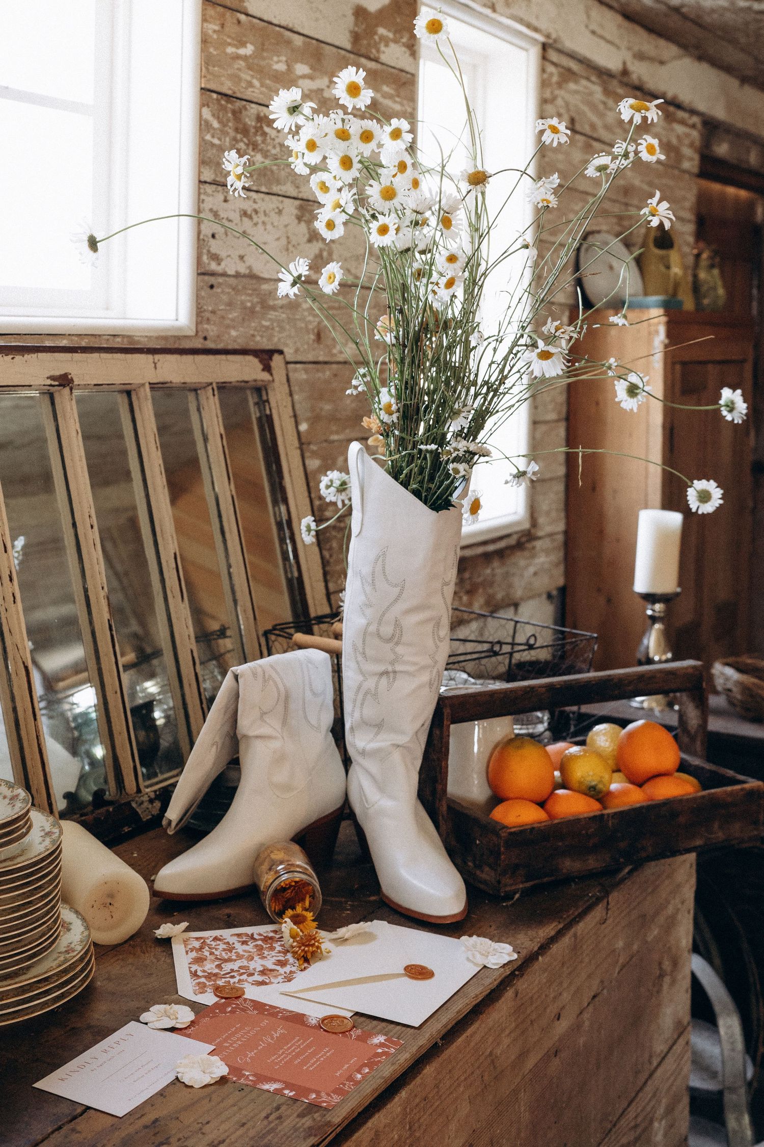 White cowboy boots used as vase for daisies beside rustic wooden crate with citrus.