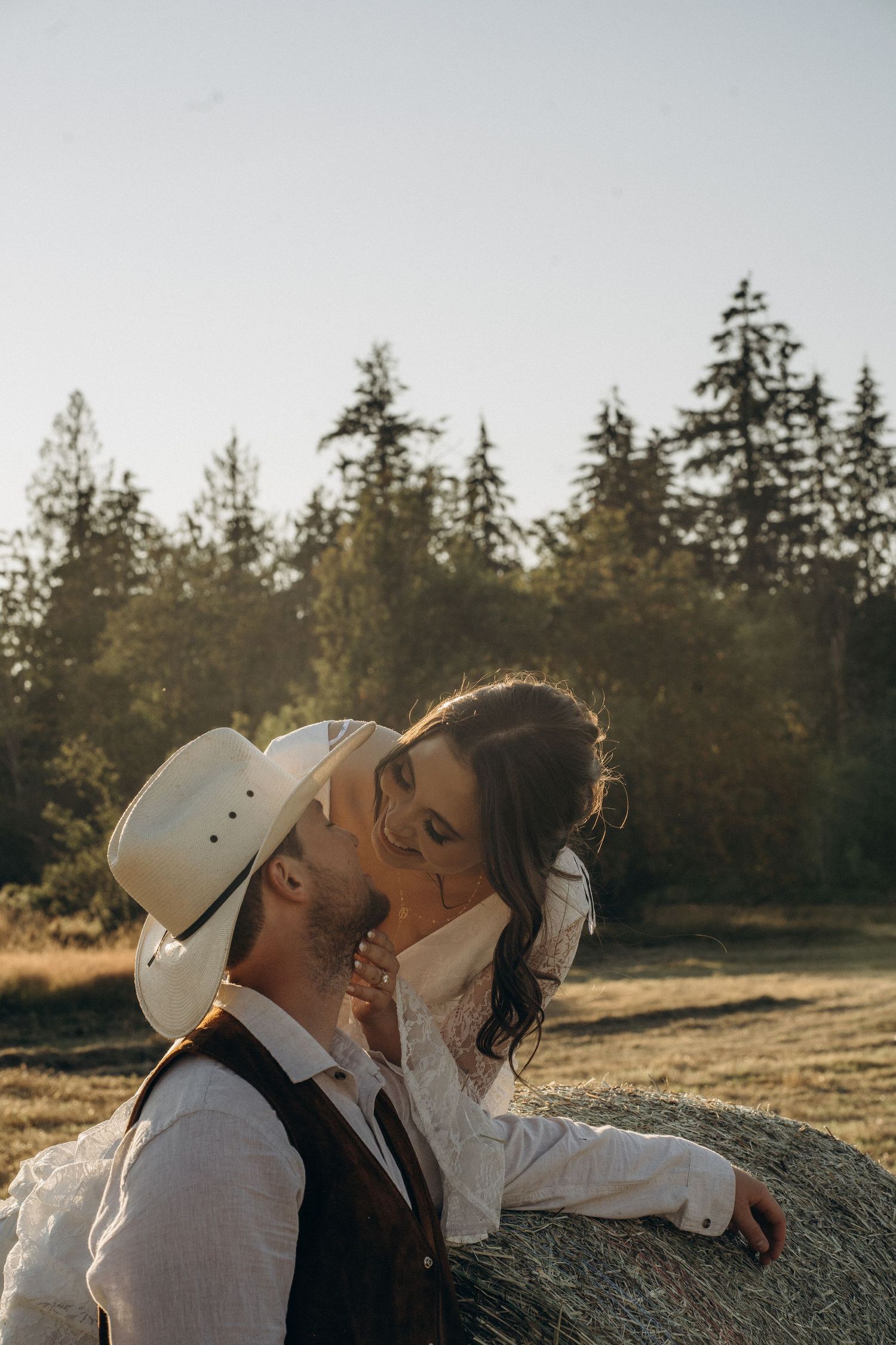 Bride and groom share a romantic kiss by a hay bale at golden hour in a rustic country field.