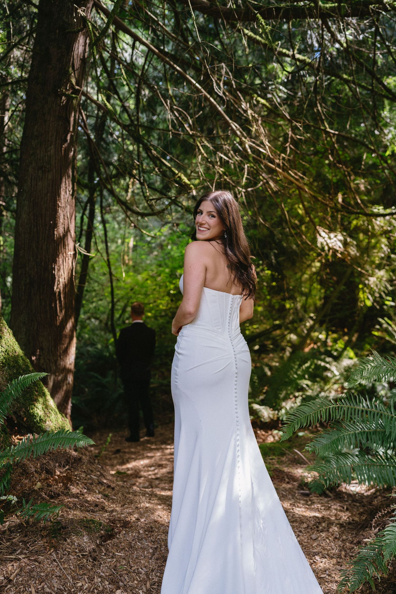 Bride in strapless white gown smiles over shoulder on forest path.