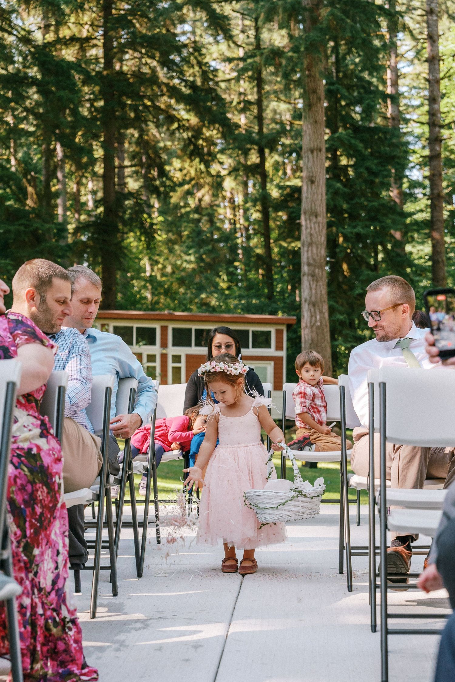 Flower girl in pink dress scatters petals down wedding aisle between seated guests in forest venue.