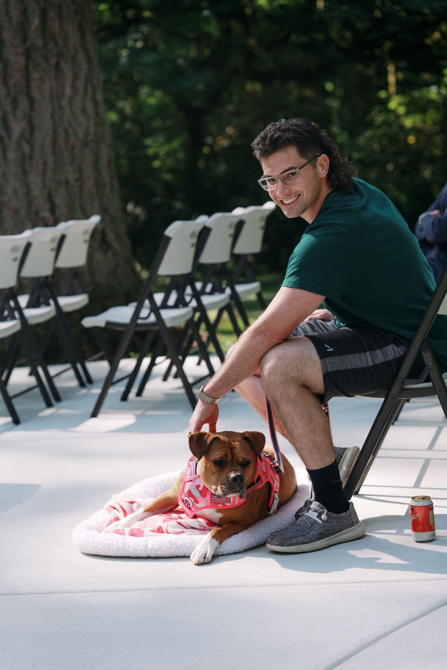 Man in green shirt petting a dog dressed in pink outfit on a white pet bed at an outdoor wedding venue.