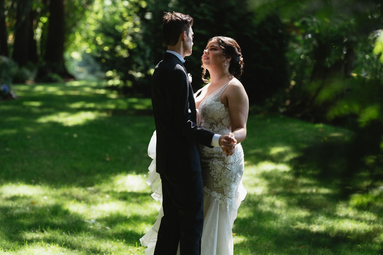 Bride in lace gown and groom in black tuxedo share an intimate moment on a shaded green lawn.