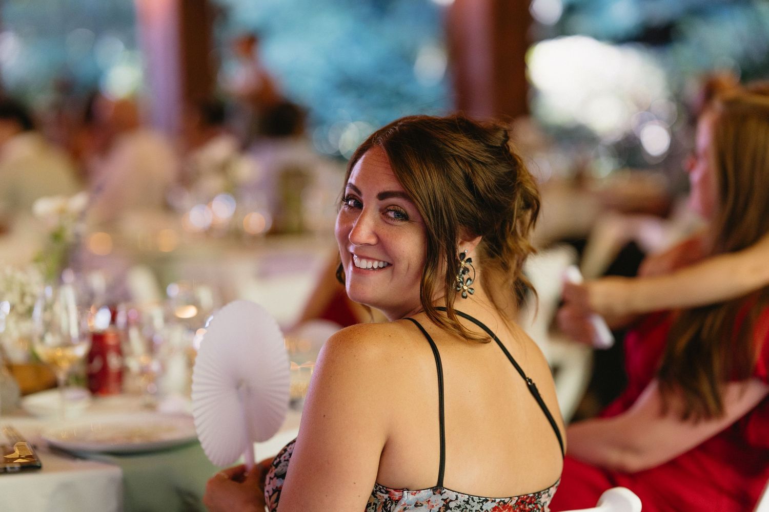 Smiling wedding guest in sparkly dress holds fan at elegant outdoor reception dinner.