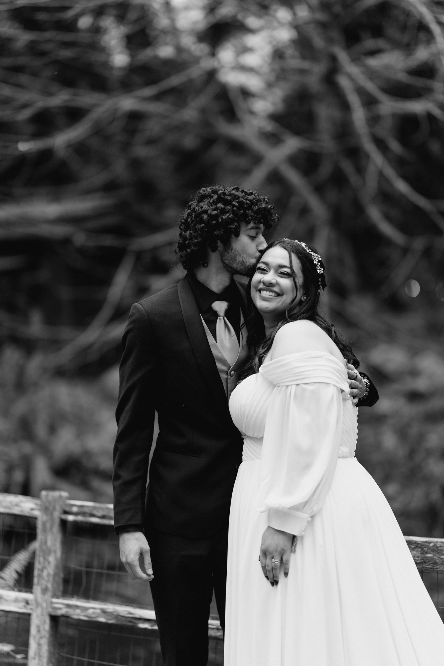 Groom kisses bride on cheek as she smiles in elegant black and white wedding portrait.