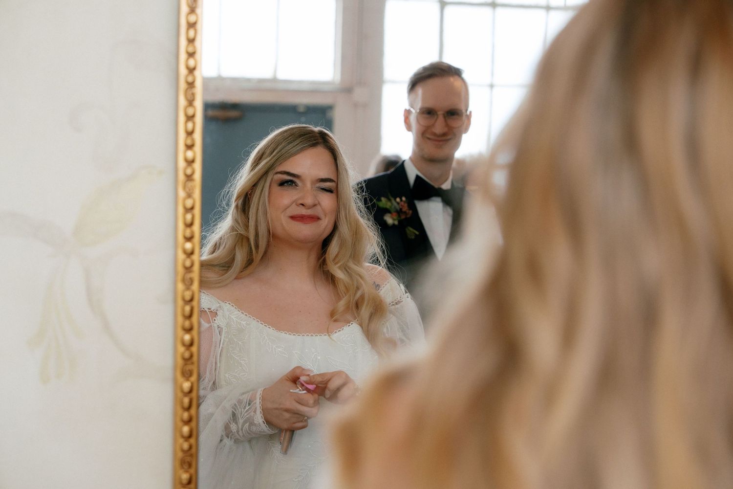Bride and groom smiling at their reflection in ornate gold-framed mirror on wedding day.