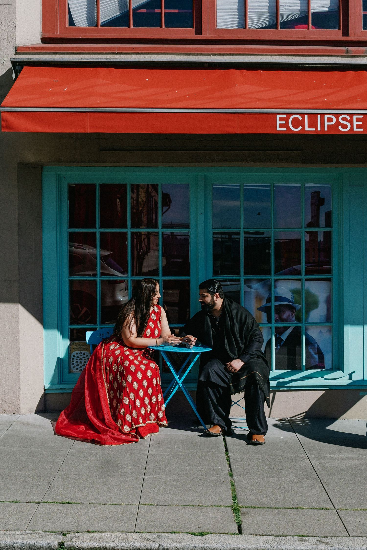 South Asian couple in traditional attire sitting at an outdoor cafe table beside a turquoise storefront.