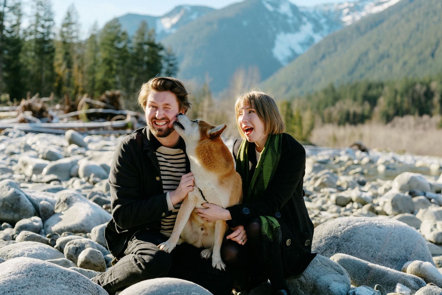 Laughing couple sitting on rocks with their Shiba Inu dog licking their faces in mountain setting.