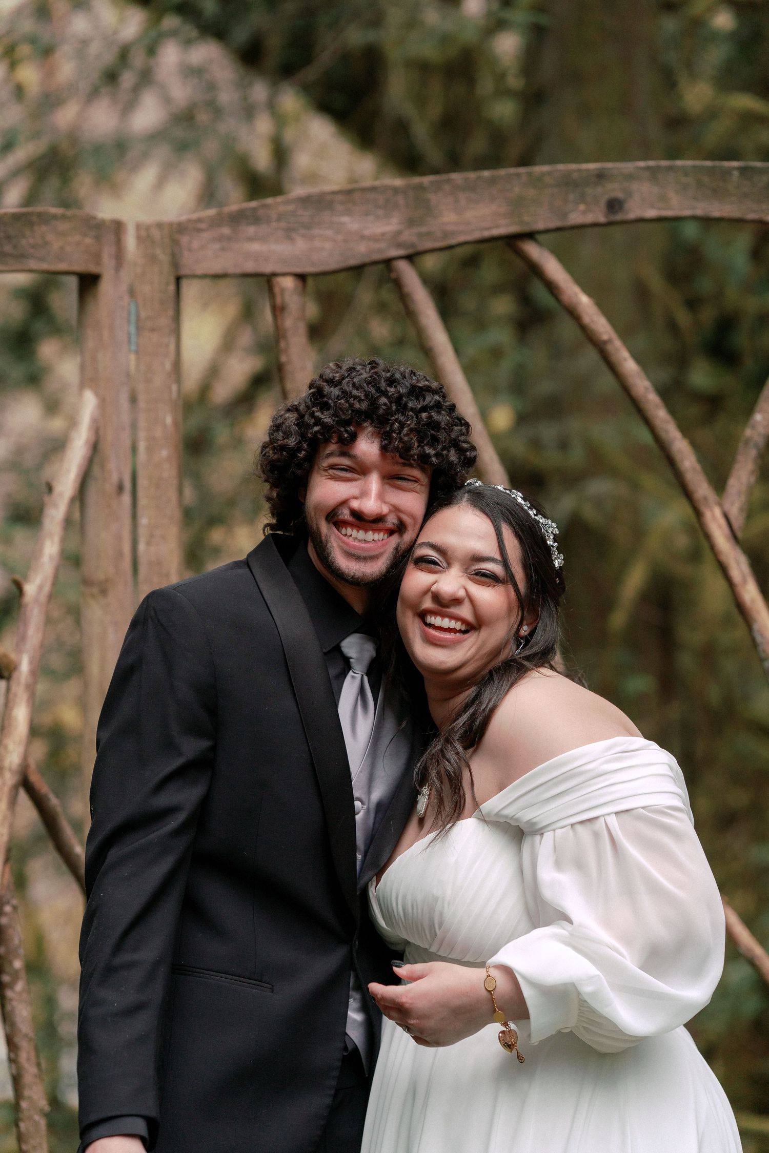 Happy newlywed couple smiling and embracing under rustic wooden arch, bride in white gown with floral crown.