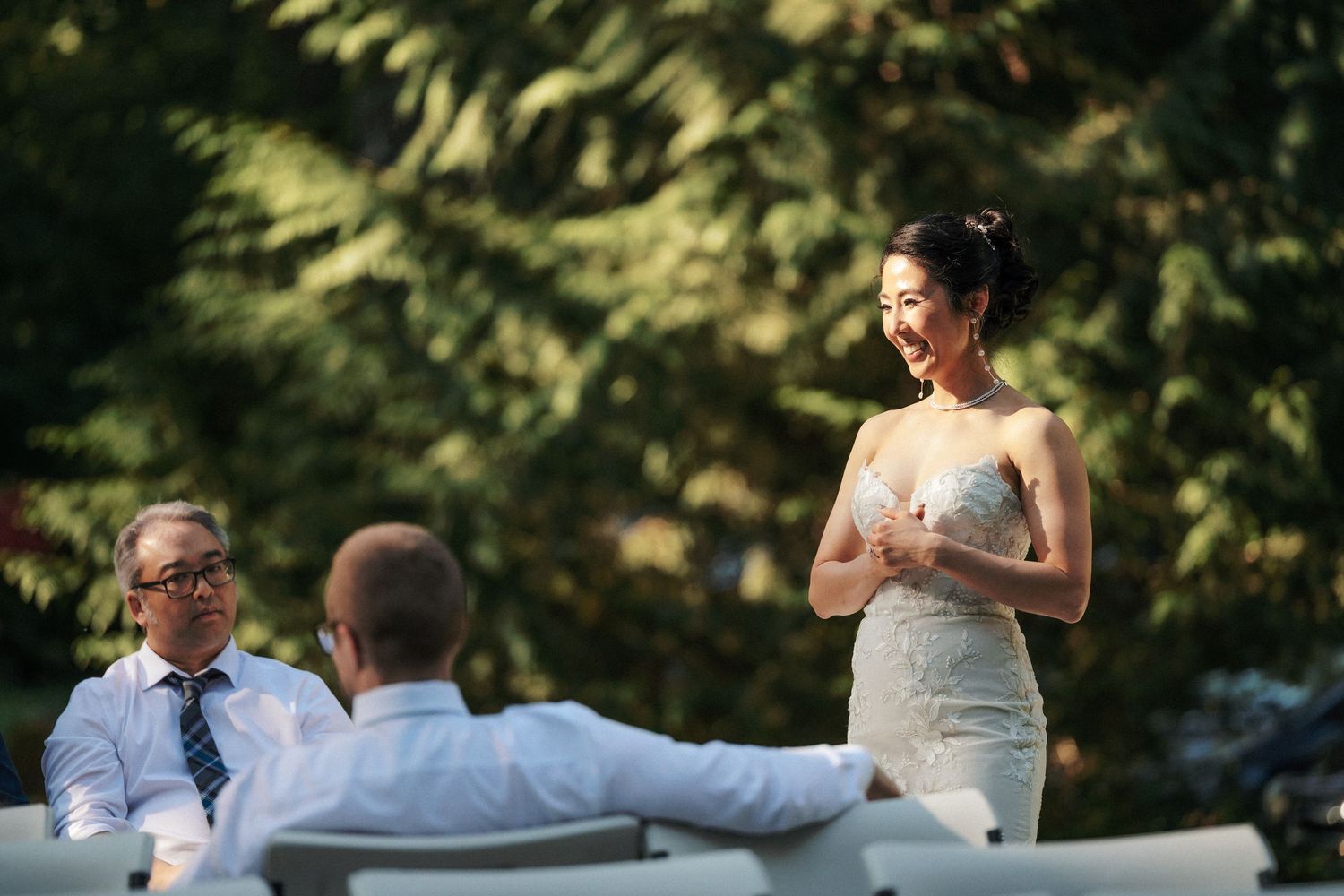 Bride in elegant strapless gown smiles while speaking at outdoor wedding reception among guests.