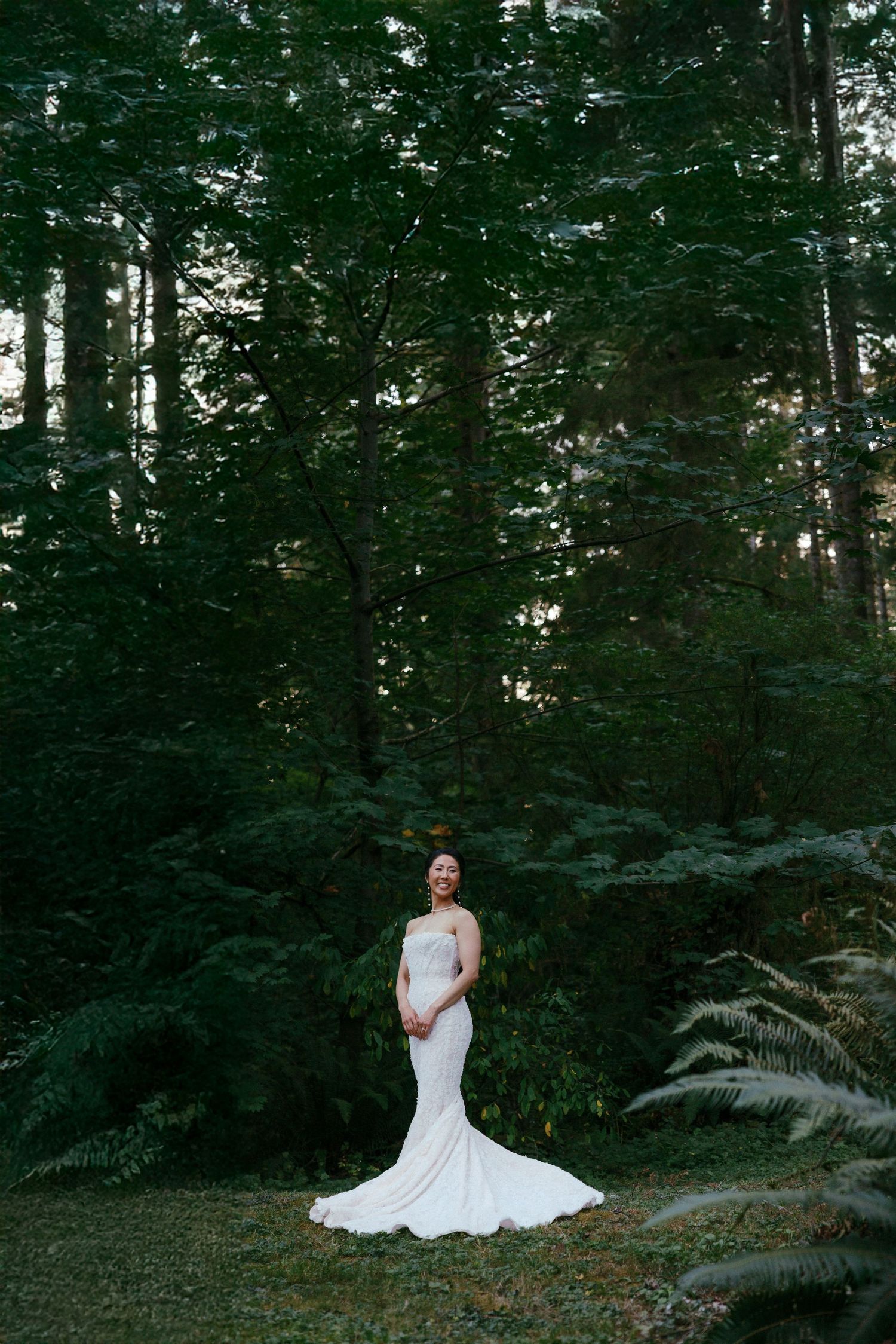 Elegant bride in fitted white mermaid gown stands alone among towering evergreen trees in a forest.
