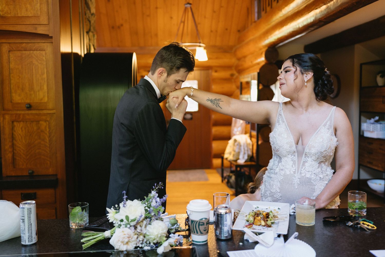 Bride feeds groom cake at rustic wooden cabin wedding reception table with white flowers.