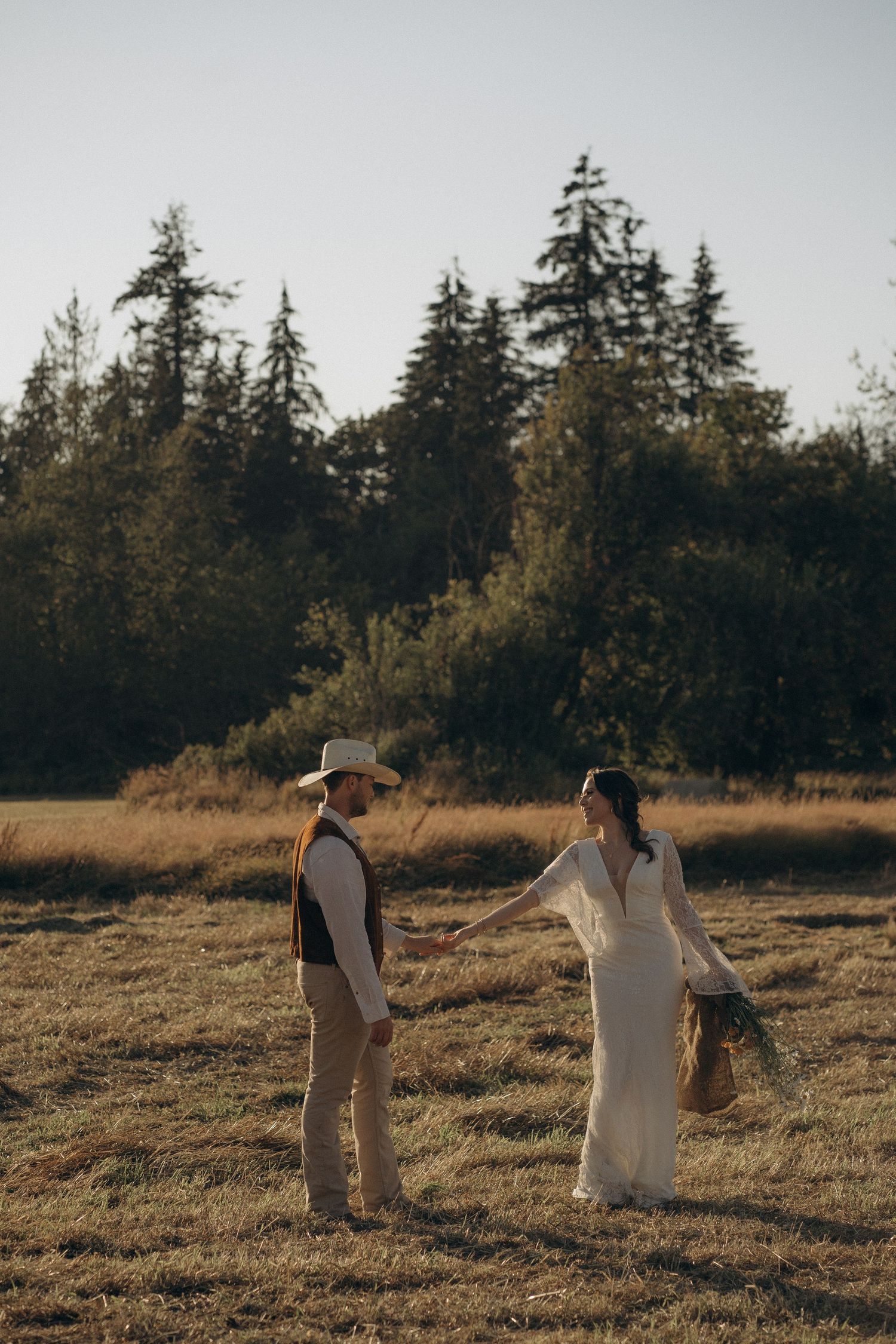 Western wedding couple holding hands walking through golden field with pine trees at sunset.