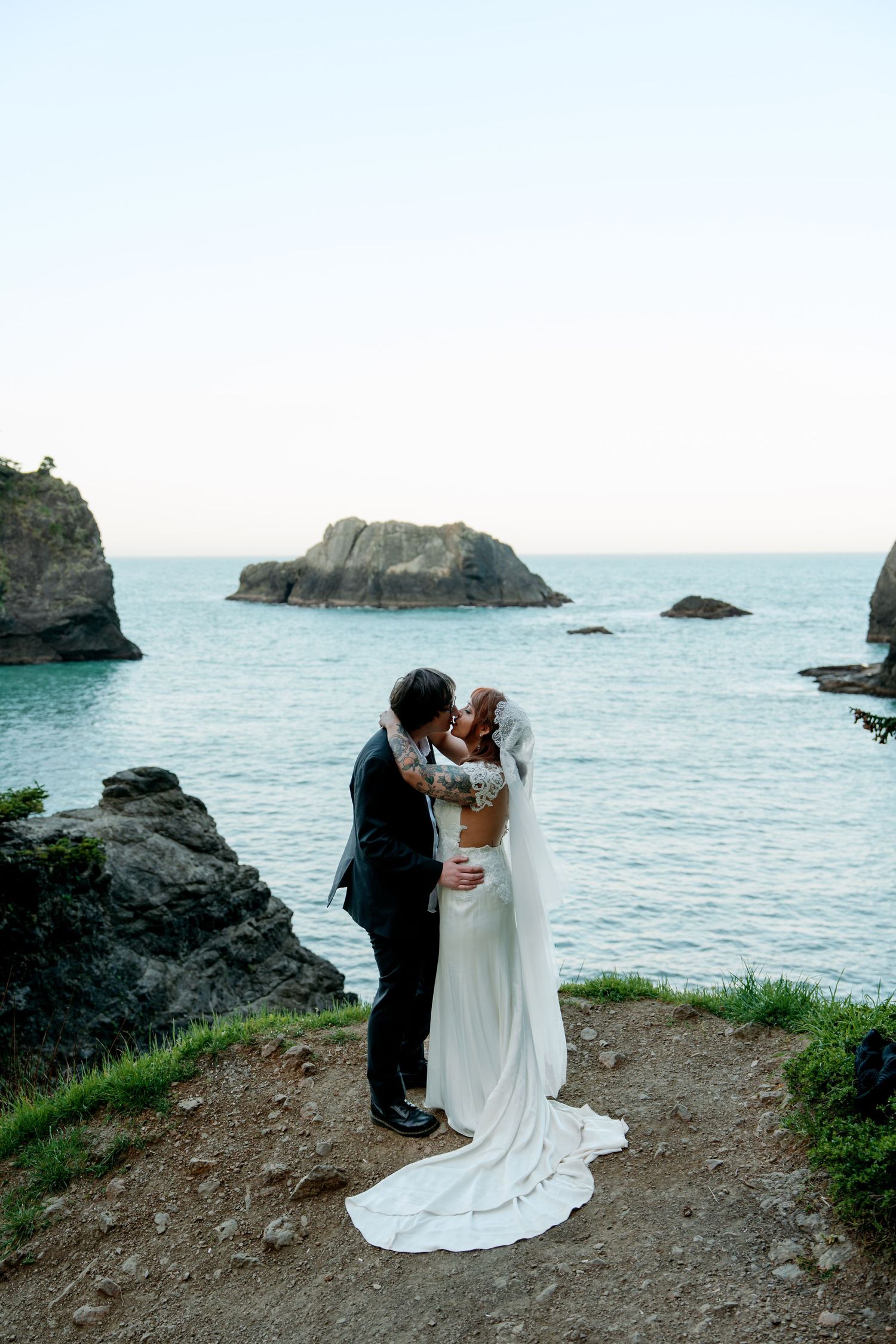 Bride and groom share a romantic kiss on a rocky coastal cliff overlooking the ocean.