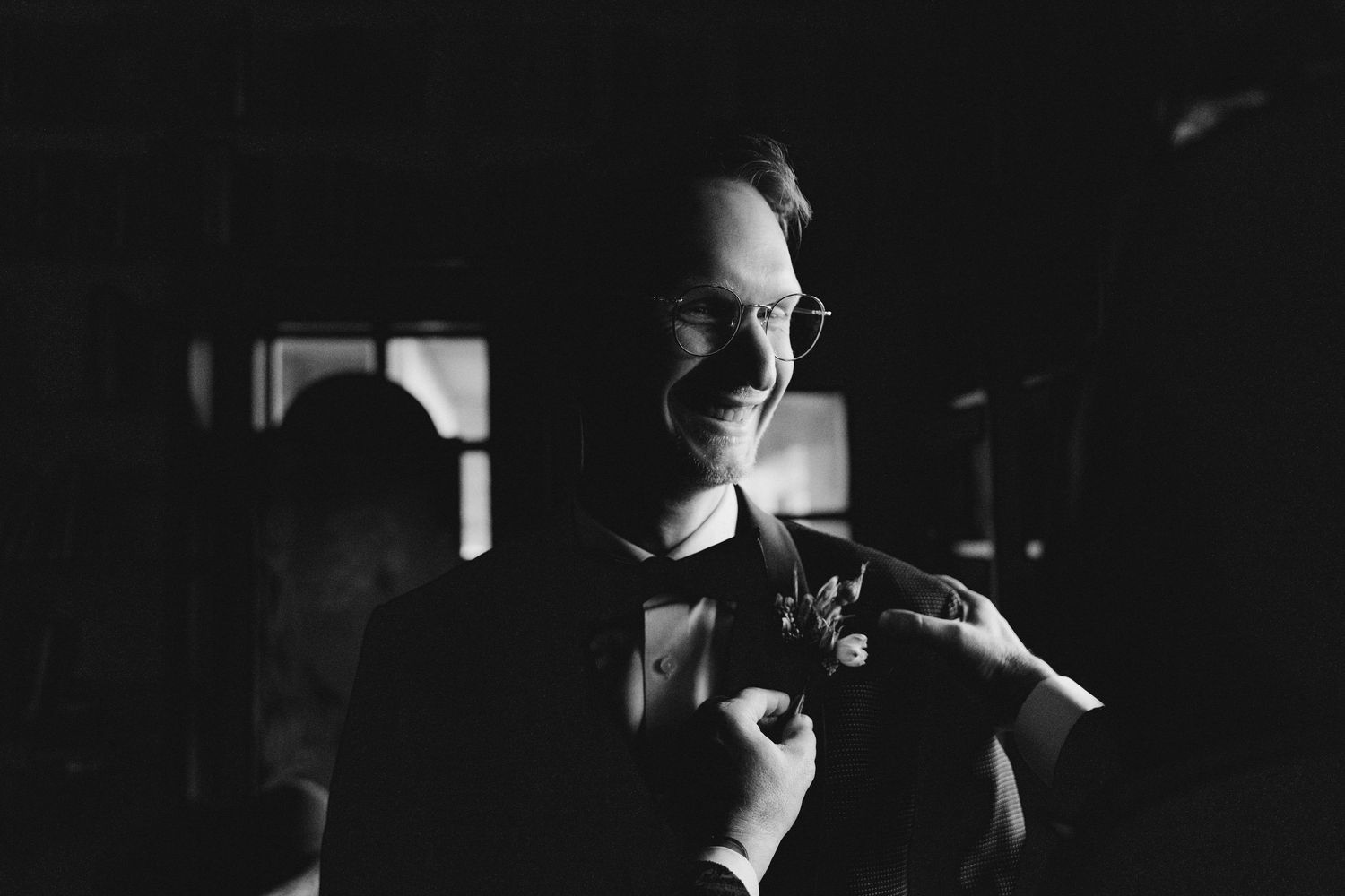Black and white portrait of groom in tuxedo smiling while boutonniere is pinned on lapel.