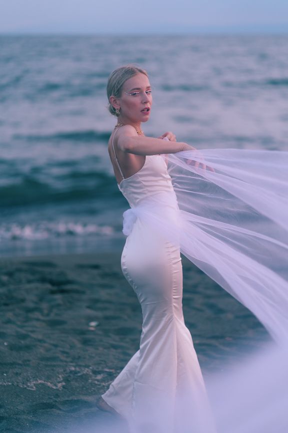 Woman in white mermaid gown releases flowing veil into ocean breeze at twilight.