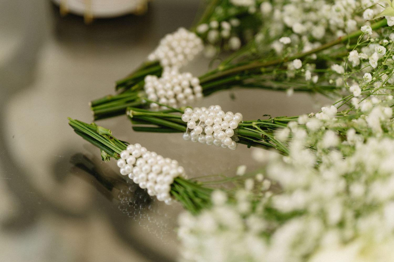 Small white pearl-wrapped baby's breath boutonnieres arranged on a reflective surface.