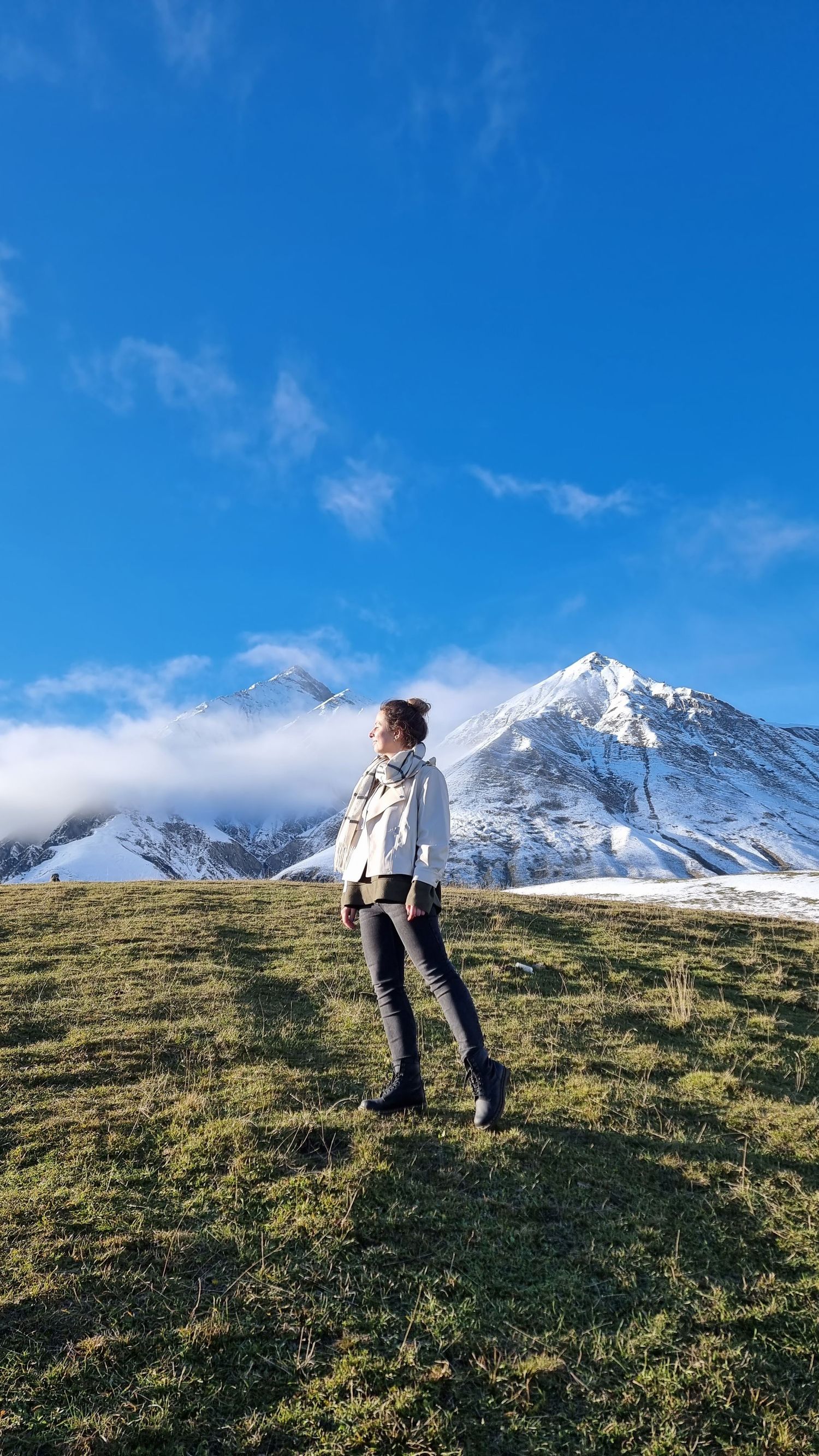 Woman in white jacket and scarf standing on green hillside with snow-capped mountain peak under blue sky.